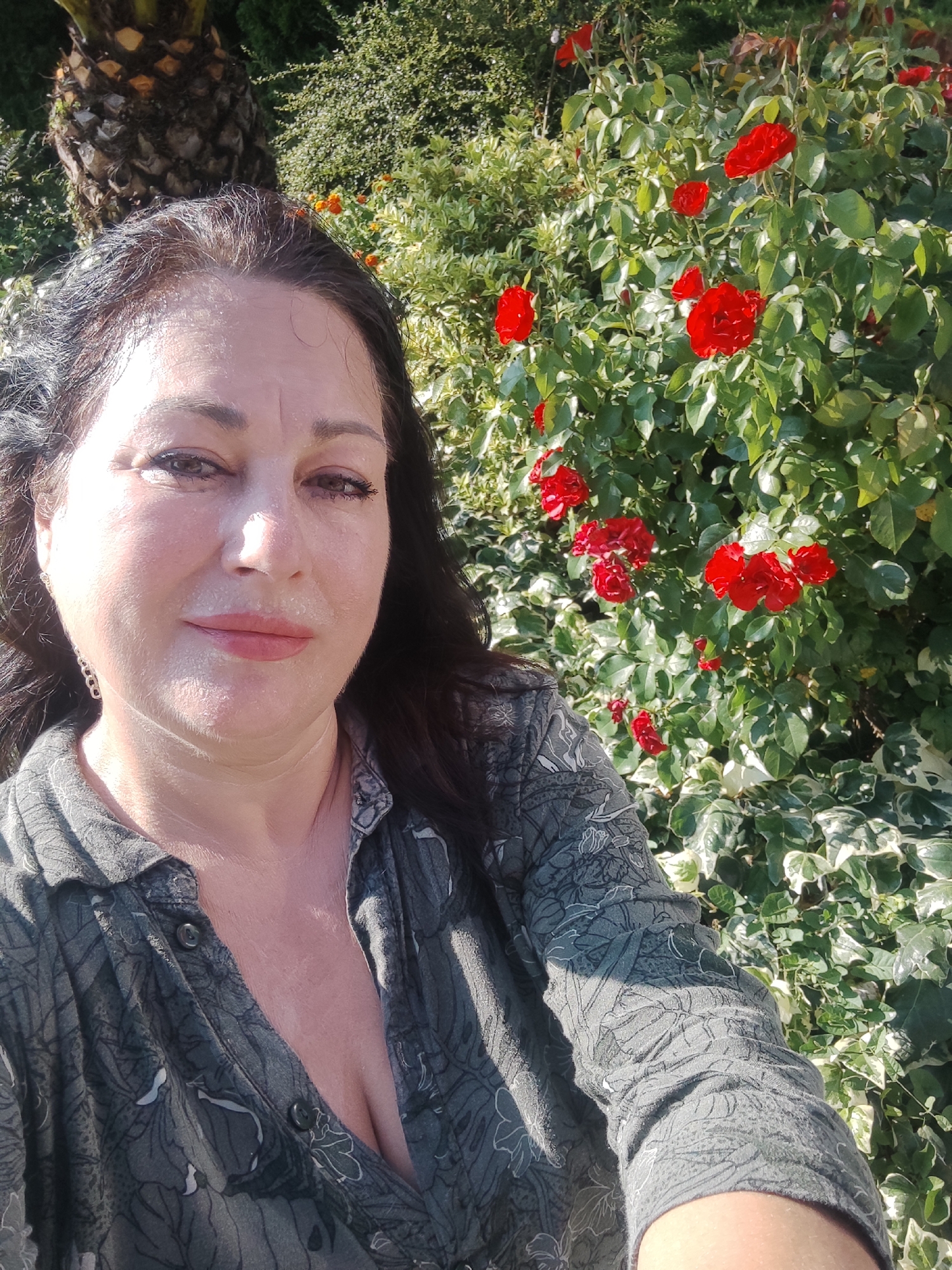 A woman with dark hair and fair skin is posing for a selfie outdoors. She is wearing a dark green patterned shirt that is unbuttoned, revealing her décolletage. The woman has make-up on, including mascara and lipstick. She is wearing an earring in her left ear. In the background, there is a lush green bush with red roses and a palm tree trunk. The lighting suggests it is daytime and sunny. The image appears to be taken in a garden or park setting. There is no indication in the image of the specific location being the city of Sochi, Russia.