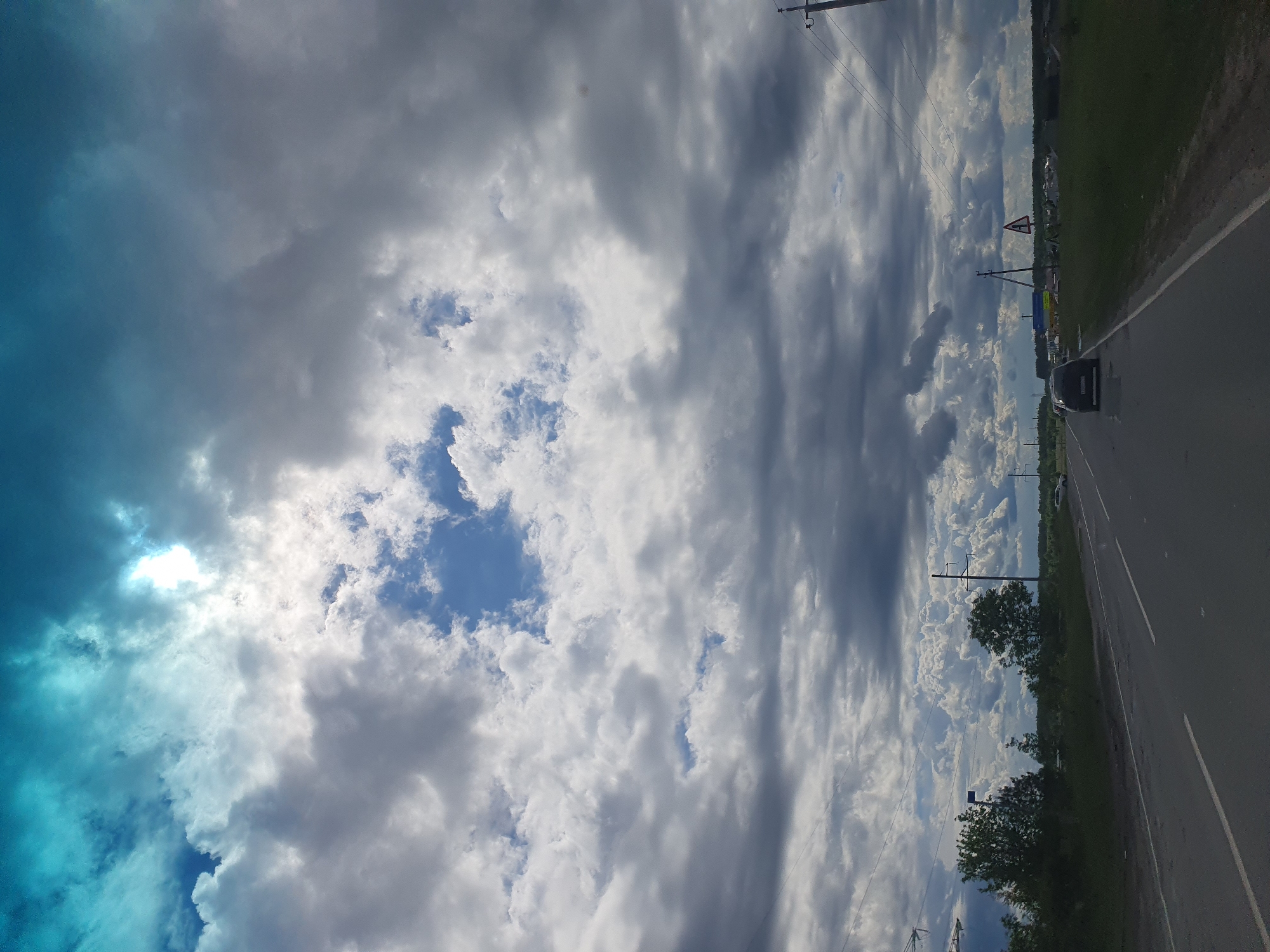 The image depicts a road in what appears to be a rural area, likely near Архангельское, Russia, given the prompt. The road surface is asphalt and includes a vehicle driving on the road. Overhead power lines are present along the side of the road. The sky is filled with a mix of blue sky and dense clouds, some appearing white and others dark gray, potentially indicative of an approaching storm. Vegetation, likely grass and trees, lines the edge of the road. A road sign is visible alongside the road. The image appears to have been taken from within a moving vehicle.