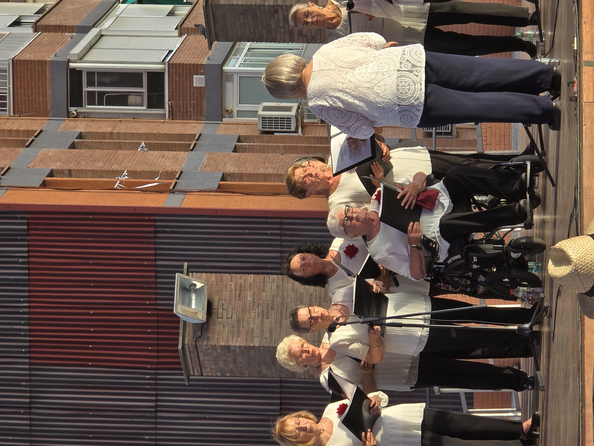 A group of women, some standing and one seated in a wheelchair, are gathered on a wooden stage, performing as a choir. They are all dressed in white tops and black pants. The woman in the wheelchair is positioned towards the front and center of the group. Behind them, the corrugated metal facade of a building is visible, with a section painted red. To the right, a brick building with windows and balconies is visible. The scene appears to be an outdoor performance, likely during daylight hours. This performance is taking place in Cornellà de Llobregat, Spain.