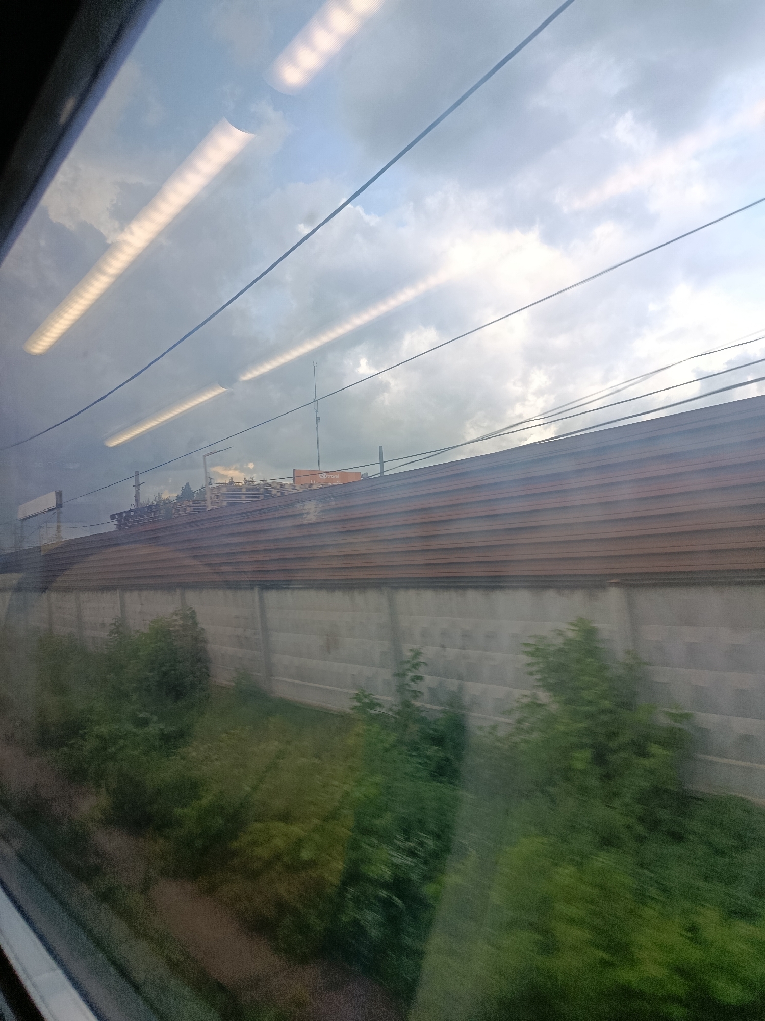 The image shows a view from a moving vehicle, likely a train, looking out a window. The scene is dominated by a long, horizontal, brown-tiled structure that runs across the middle ground. Below this structure is a gray, concrete-block wall, with patches of green vegetation growing in front of it and along the ground. Above the tiled structure, a slightly blurry industrial or commercial area with buildings and a tall antenna is visible against a cloudy sky. Several thin, dark lines, likely overhead wires, cross the sky. The reflection of fluorescent lights from inside the vehicle is visible in the upper portion of the image. The image appears to have been taken in Elektrougli, Russia, based on the context provided.