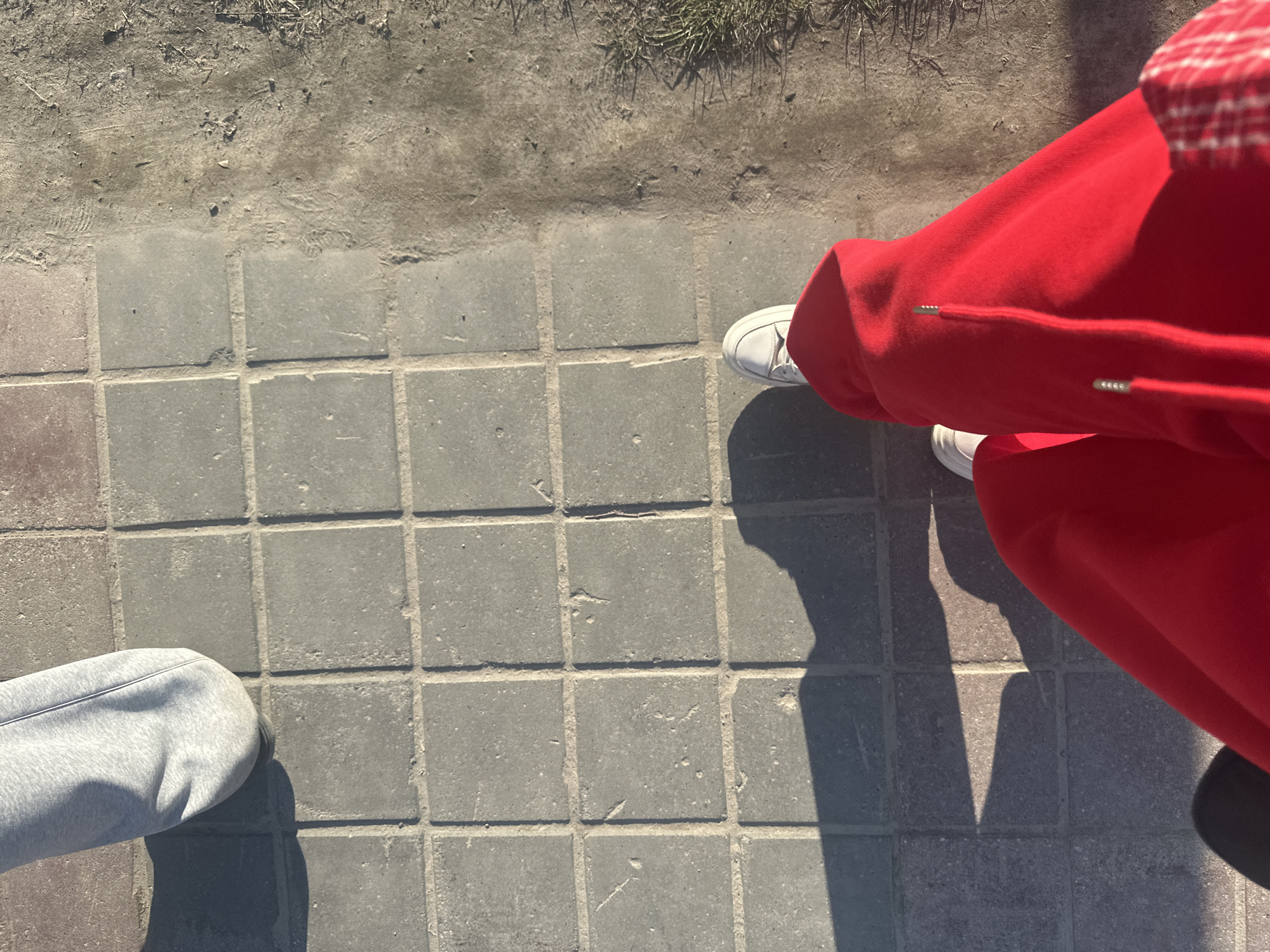 The image shows a street scene from a high-angle perspective. Two individuals are partially visible, one wearing grey sweatpants and white sneakers, the other wearing red sweatpants and white sneakers. They are standing on a paved area with square grey stones. To the left, there is a dirt area with some sparse grass. Strong shadows cast by the individuals are visible on the pavement. The location is not specified as Megion, Russia.