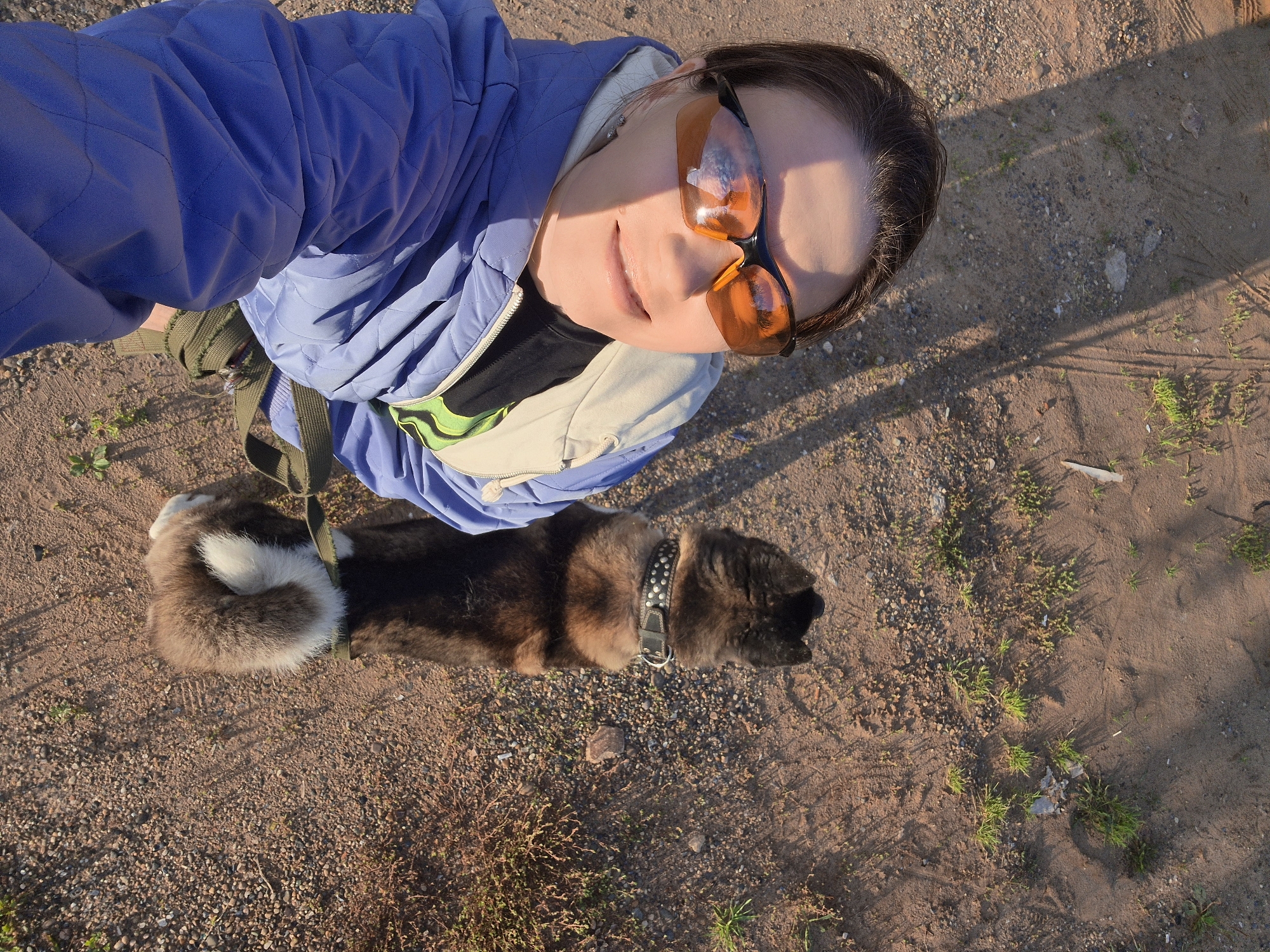 A woman wearing orange-tinted sunglasses and a blue quilted jacket stands on a dirt path in Chaikovsky, Russia, taking a selfie. She is accompanied by a medium-sized dog with brown and black fur, wearing a studded collar. The dog is facing away from the camera, looking towards the right. The ground is covered with small stones and sparse dry grass, with shadows from an unseen structure falling across the path. The overall lighting suggests it is daytime, possibly late afternoon.