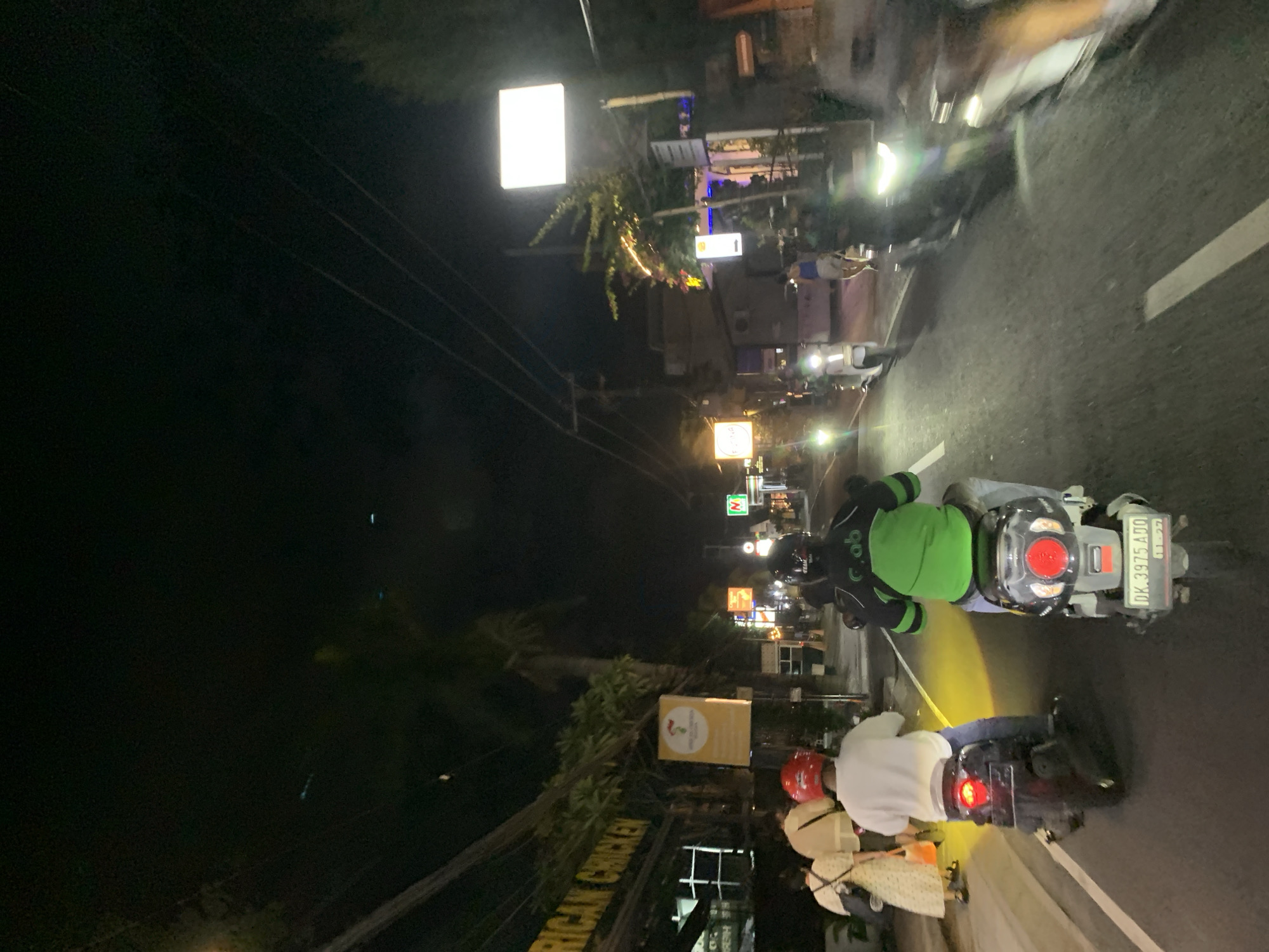 At night in North Kuta, Indonesia, the scene captures two scooters on a road. One scooter is operated by a person wearing a green jacket and a black helmet; this scooter has a license plate of "DK 3975 ADO". The second scooter has three people on it: a driver wearing a red helmet and two passengers. Streetlights illuminate the area, highlighting the "American Chicken" storefront and other businesses.