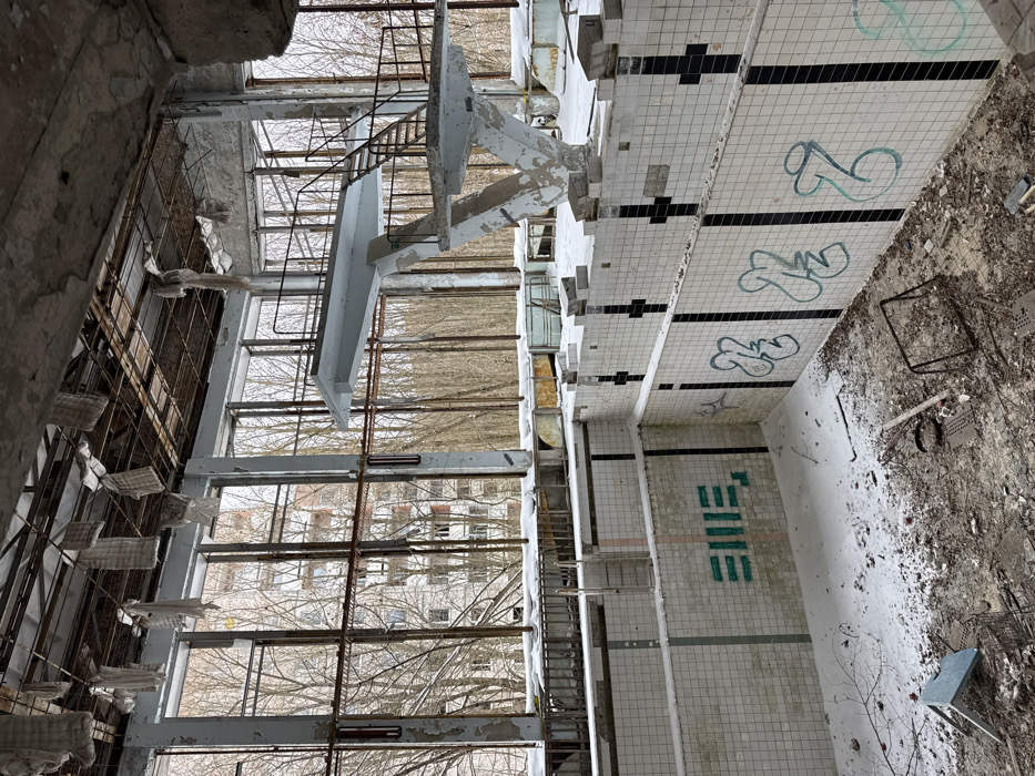 The image depicts an abandoned and heavily decayed indoor swimming pool, captured from an elevated perspective looking down into the pool area. The photograph appears to be rotated 90 degrees counter-clockwise.
The central feature is the empty swimming pool, tiled with a grid pattern of white and black squares. Black lane markers are visible on the pool's floor and walls, with several smaller, black square markings along the top edge of the pool. The bottom of the pool and the surrounding floor are covered in dirt, debris, and what appears to be dust or light snow.
A prominent diving platform, constructed from what looks like concrete, stands at one end of the pool. It has a set of metal stairs leading up to it, both showing significant signs of wear, peeling paint, and rust. The platform itself appears sturdy but weathered.
Along one side of the building, large, tall windows span from floor to ceiling, revealing an exterior scene. Through these windows, a landscape of bare, leafless trees is visible, suggesting a winter or early spring season. Beyond the trees, another large, dilapidated building, possibly an apartment block, can be seen, reinforcing the impression of an abandoned urban environment. The window frames are metallic and appear corroded, with broken or missing panes in some sections.
The interior structure of the building is exposed, with rusty metal beams, supports, and remnants of what might have been wall coverings or insulation hanging from above. The overall condition of the scene indicates prolonged abandonment and neglect, with extensive decay, crumbling surfaces, and accumulated detritus throughout. Various pieces of debris, including what looks like a metal chair frame and other miscellaneous objects, are scattered on the floor around the pool.
There is no activity taking place; the scene is static and reflects a state of disuse and ruin. The atmosphere is desolate and eerie.
Visible text and graffiti include:
- On the tiled wall at the deep end of the pool, a word or set of characters in a stylized, blocky green font is painted. It strongly resembles "FIVE" or possibly the Cyrillic word "ПЯТЬ" (Pyat), which also means "five."
- Several instances of light blue or teal graffiti, appearing as abstract designs or stylized letters, are spray-painted on the white tiles along the walls of the pool. These do not form discernible words.