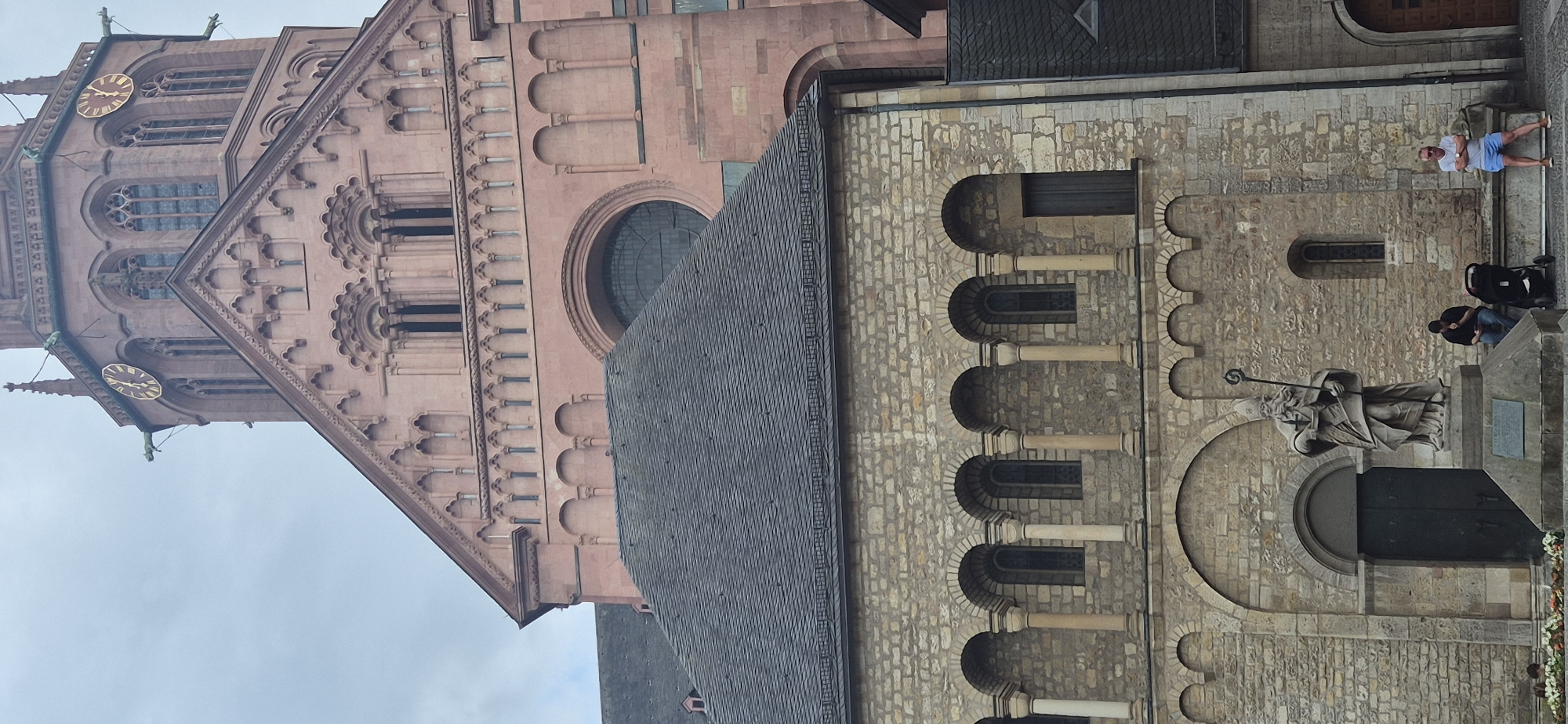 This media file depicts a view of the Mainz Cathedral in Mainz, Germany. The cathedral's exterior is visible, showcasing its distinctive red sandstone and stone masonry. The image is oriented vertically, with the cathedral's tower and rooflines dominating the frame. On the right side of the image, a stone statue of a robed figure holding a staff stands prominently. Several people are present in front of the cathedral. One person, wearing a white t-shirt and blue shorts, stands on a raised platform and appears to be looking towards the camera. Nearby, two other individuals are seated. Two strollers are also visible, suggesting families are present. The sky appears overcast, indicating a cloudy day. The architectural details of the cathedral, including its arched windows and decorative stonework, are clearly visible. The overall scene suggests a peaceful day at a significant historical landmark. No visible text is discernable in the image.