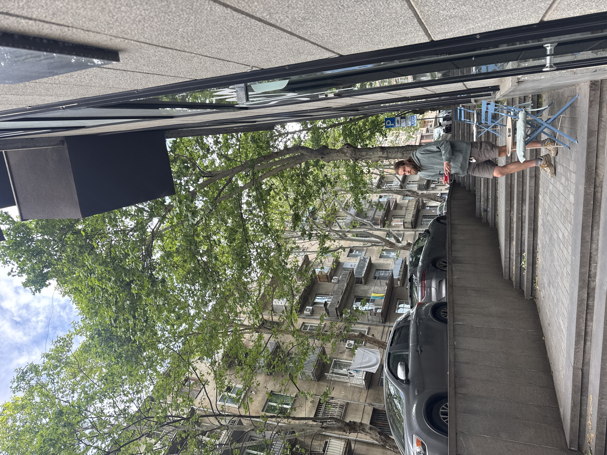 A man stands on a sidewalk with a building and cars behind him. He holds a red object in his hand. There are blue tables and chairs near him. Trees are in the scene. The location is an urban area, likely a city street.
