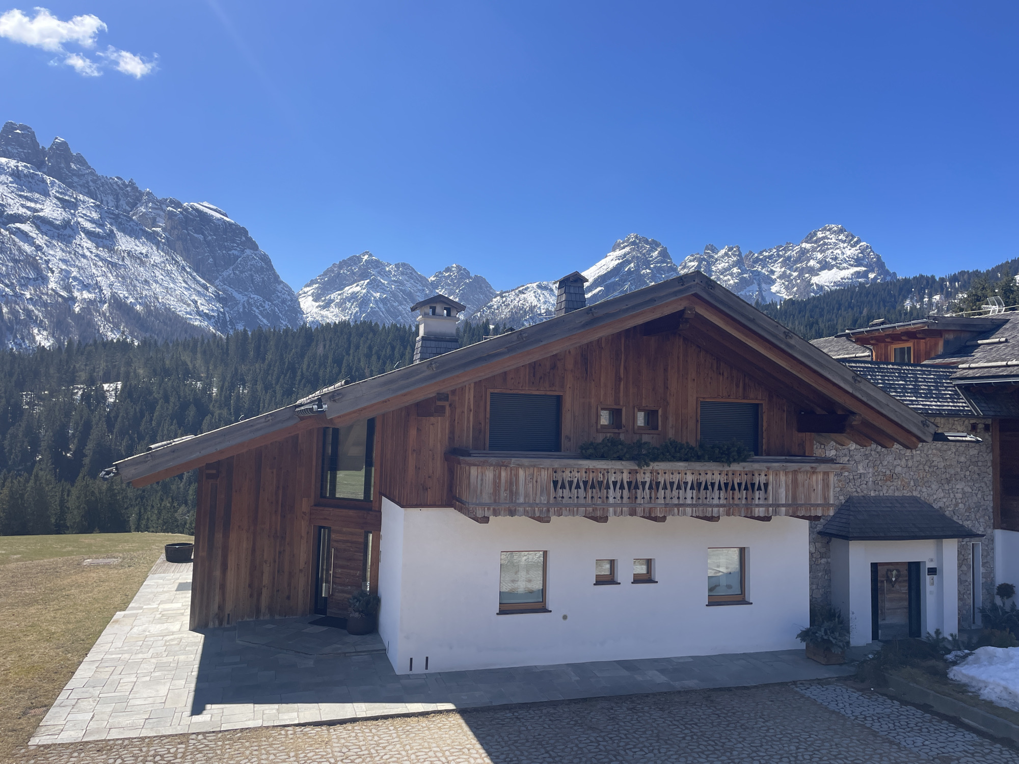 The image depicts a chalet-style house in a mountainous region, likely Sappada, Italy. The house has a distinctive architectural style with a wooden upper story and a white stucco lower story. There is a balcony with wooden railings, and some greenery is visible on the balcony. Windows are visible on both levels, some with closed shutters. The house has a sharply angled roof, and a chimney is present.
In the background, there are snow-capped mountain peaks and a dense forest of evergreen trees. The sky is clear blue with a few scattered clouds, suggesting a sunny day. The foreground shows a paved area leading up to the house. To the right, part of another building with a stone exterior is visible, along with some melting snow patches. The overall scene is characteristic of a mountain resort town.