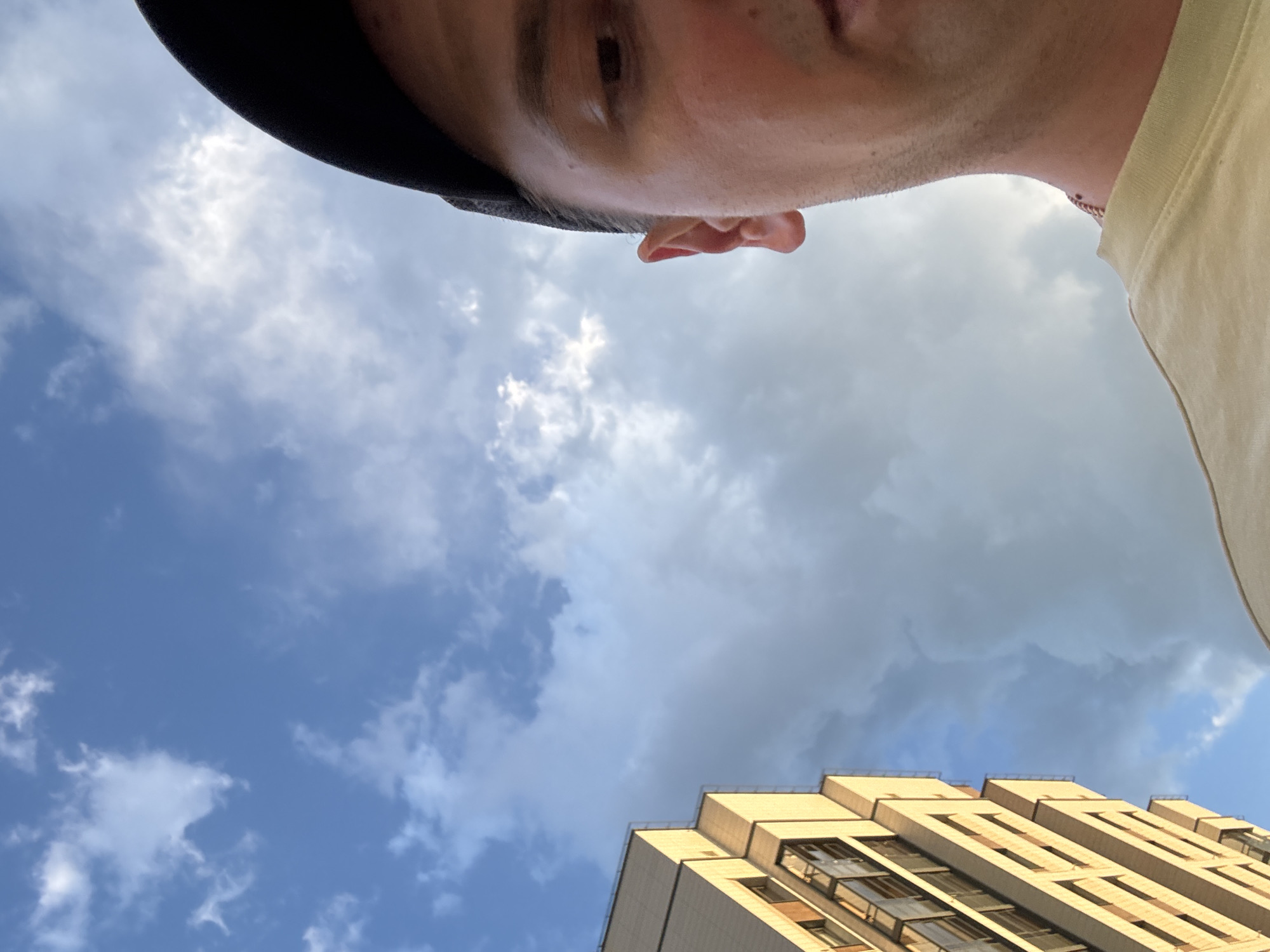 The image is a low-angle, upward-facing shot, likely a selfie. It features the upper part of a person's face, including an eye and ear, framed by a dark baseball cap. The person is wearing a light-colored t-shirt. The background consists of a sky with a mix of blue and gray clouds, suggesting a day with variable weather. A section of a modern building with light-colored facade and numerous windows is visible in the lower right portion of the frame, partially obscured by the person's t-shirt. The overall impression is casual, possibly taken outdoors on a cloudy but bright day. There is no visible text in the image. The location is identified as Koltsovo, Russia.