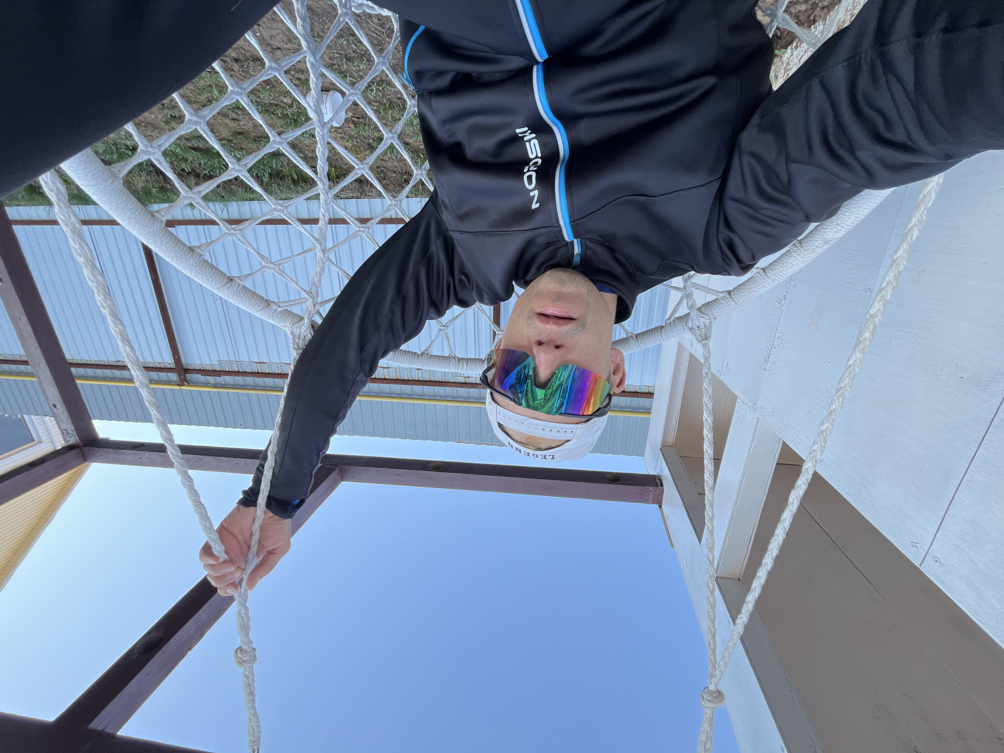 A person wearing a black jacket with blue stripes, rainbow-colored sunglasses, and a white baseball cap is hanging upside down in a rope hammock. They are holding onto the rope of the hammock with their right hand. The background features a blue sky, a corrugated metal roof, a section of white netting, and wooden structural supports painted white and brown. The location is Chaikovsky, Russia.