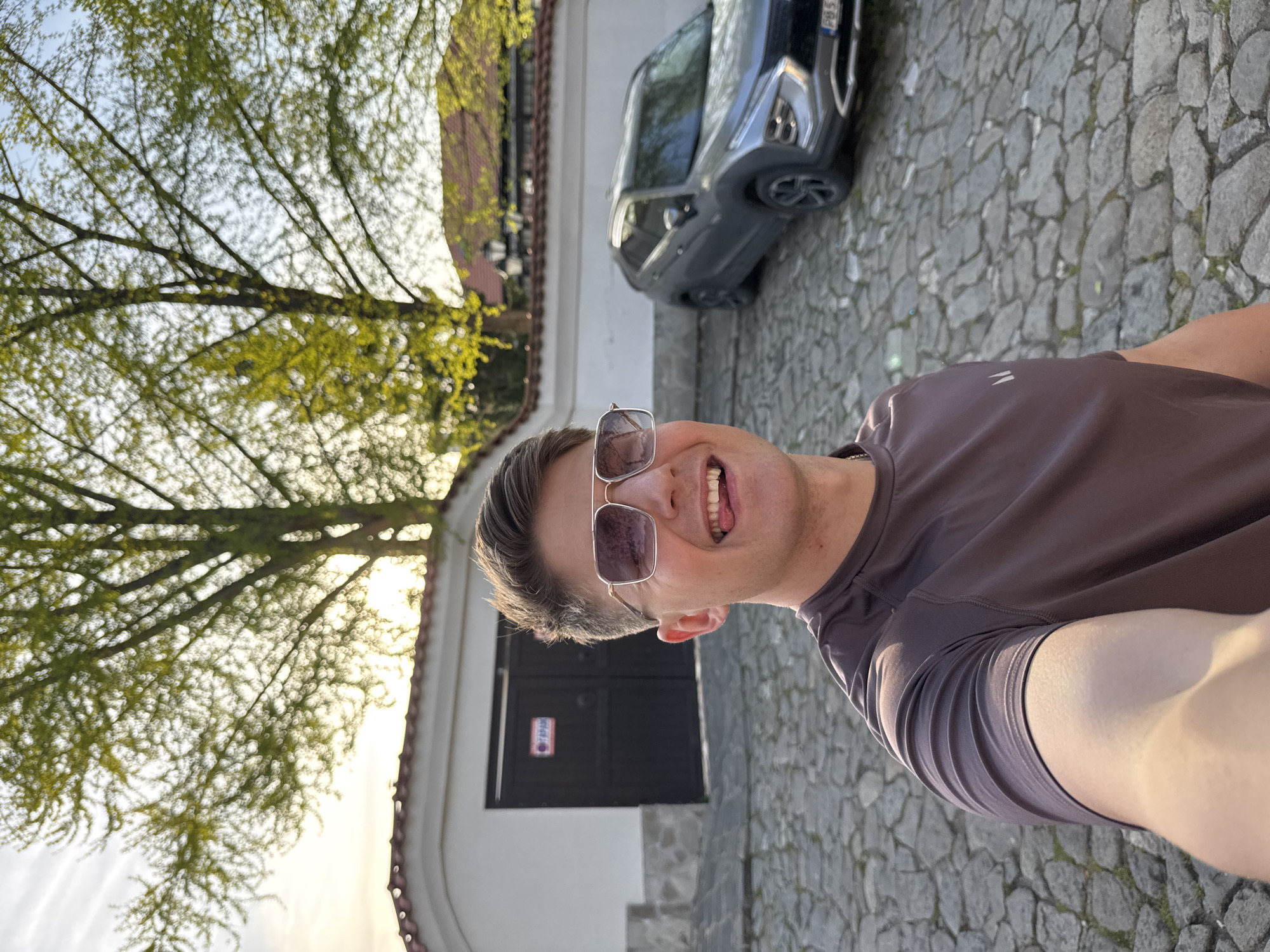 A close-up photograph shows a man in sunglasses taking a selfie. He is wearing a brown t-shirt. Behind him is a white building with a dark gray door and a gray car. A tree with light green leaves is to the left of the building. The ground is paved with cobblestones. This appears to be in Plovdiv, Bulgaria.