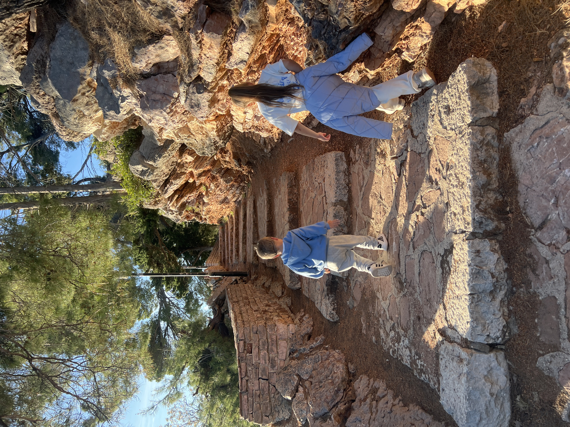 The image captures a scene on a stone path or stairway in Sveti Stefan, Montenegro. The path is constructed from large, irregularly shaped stones and bordered by rock formations and vegetation. A young woman in a light blue quilted jacket and white pants stands near the top of the stairs, while a young boy in a blue hooded sweatshirt and gray pants is further down the path. The environment includes trees and other greenery, with a glimpse of the sky visible through the foliage. The lighting suggests it is a sunny day.