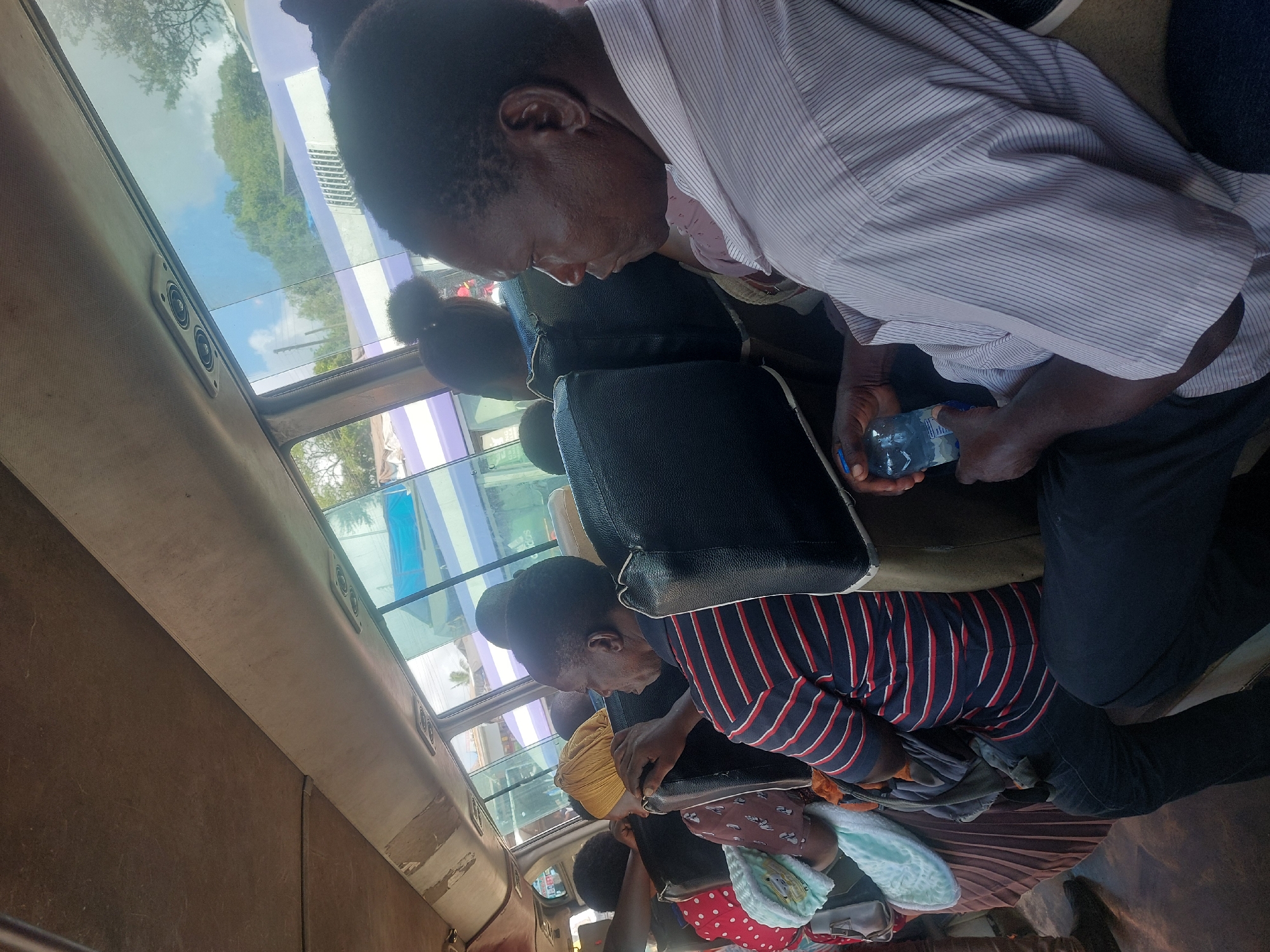 The image captures the interior of a public transport vehicle, likely a bus or minibus, in Dar es-Salaam, Tanzania, during daylight hours under a clear, sunny sky. The perspective is from slightly above and behind the passengers, looking forward and across the aisle.
Several passengers are visible seated in dark, possibly faux leather, seats. In the foreground, an adult wearing a light-colored striped collared shirt is seated, seen in profile, holding a clear plastic water bottle in both hands. Their legs, clad in dark trousers, are visible below. Behind this individual, the back of another passenger's head is visible, resting against a seat. Further into the vehicle, another adult, wearing a dark and red-striped shirt, is seated. Below them, a small child or baby, dressed in a red polka-dot garment and partially covered by a light blue patterned blanket or towel, is visible, likely on an adult's lap. Another person with a yellow head covering is visible in the distance.
The vehicle's interior features large windows along the side and possibly an overhead skylight, through which blue sky, white clouds, and green trees are clearly visible. Structures or buildings with white and purple elements can also be seen outside. The ceiling lining is a light, worn fabric, and an overhead panel with two circular speakers or vents is present. The side walls of the bus appear to be a simple, worn brown material. There are no strong emotions discernible, and the passengers appear calm while traveling. No visible text is clear in the image.