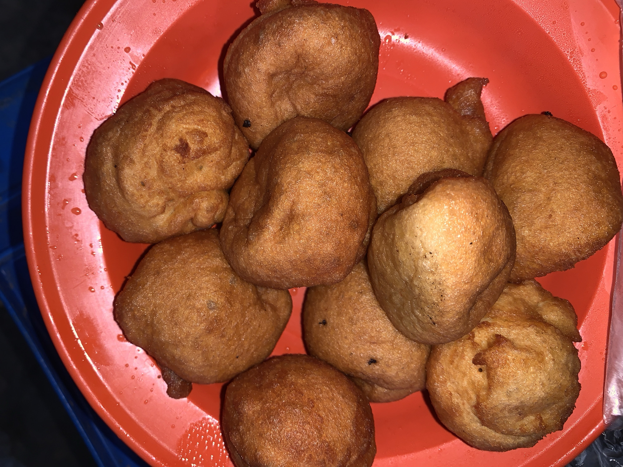 The image displays a close-up, top-down view of a serving of golden-brown, fried dough balls, commonly known as puff-puff in Nigeria. Approximately 10-12 pieces of these irregularly shaped snacks are piled within a round, red plastic bowl, occupying most of its space. The dough balls appear freshly fried, exhibiting a slightly oily sheen and textured, often crinkled surfaces. Some pieces show darker, crispier areas.
The red plastic bowl has a smooth surface with a slightly raised rim. Numerous small water droplets or condensation marks are visible across the interior of the bowl, suggesting moisture. The lighting in the scene is somewhat dim, implying an indoor setting or evening time, with artificial light sources casting soft highlights on the food and the bowl.
A hint of a blue surface, possibly a chair or table, is visible beneath the red bowl along the bottom and left edges of the frame. There are no people visible, and no specific activity is taking place other than the presentation of the food. There is no visible text in the image. The scene depicts a ready-to-eat snack, likely from a vendor or casual setting, consistent with food found around places like a stadium in Nigeria.
