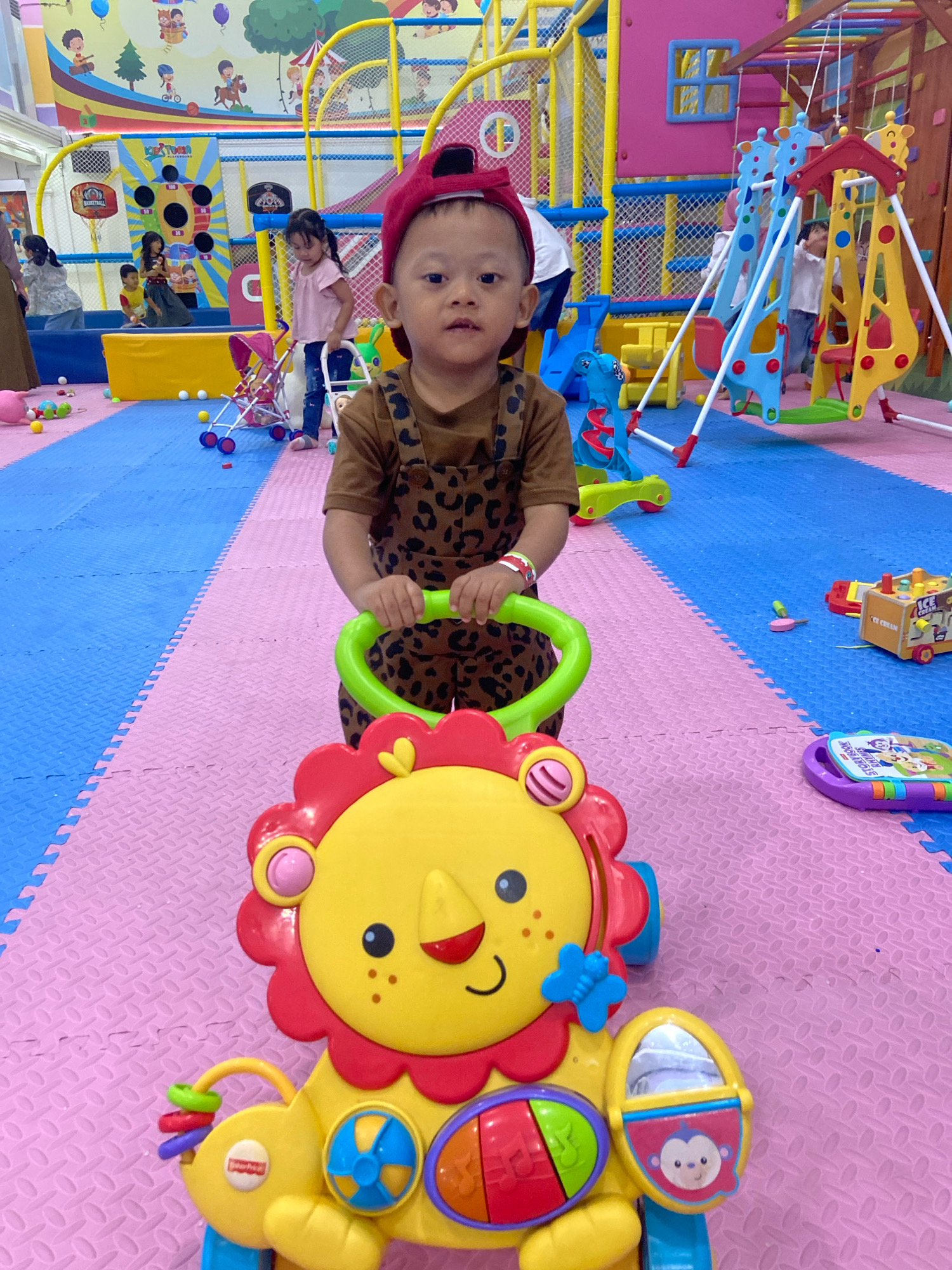 In an indoor children's play area, a young toddler wearing a brown leopard-print romper and a red baseball cap stands facing the camera, holding onto the green handles of a yellow lion-shaped push toy. The toy has a red mane, a yellow face with black dots for cheeks and a smiling mouth, and various colorful interactive elements. The toddler has a white band on their left wrist. The play area features brightly colored foam floor mats, with a blue section in the background and a pink section in the foreground. In the background, several other children are visible, along with playground equipment such as swings and climbing structures. A pink toy stroller is also present on the blue matting. The overall environment suggests a lively and safe space for children's activities.