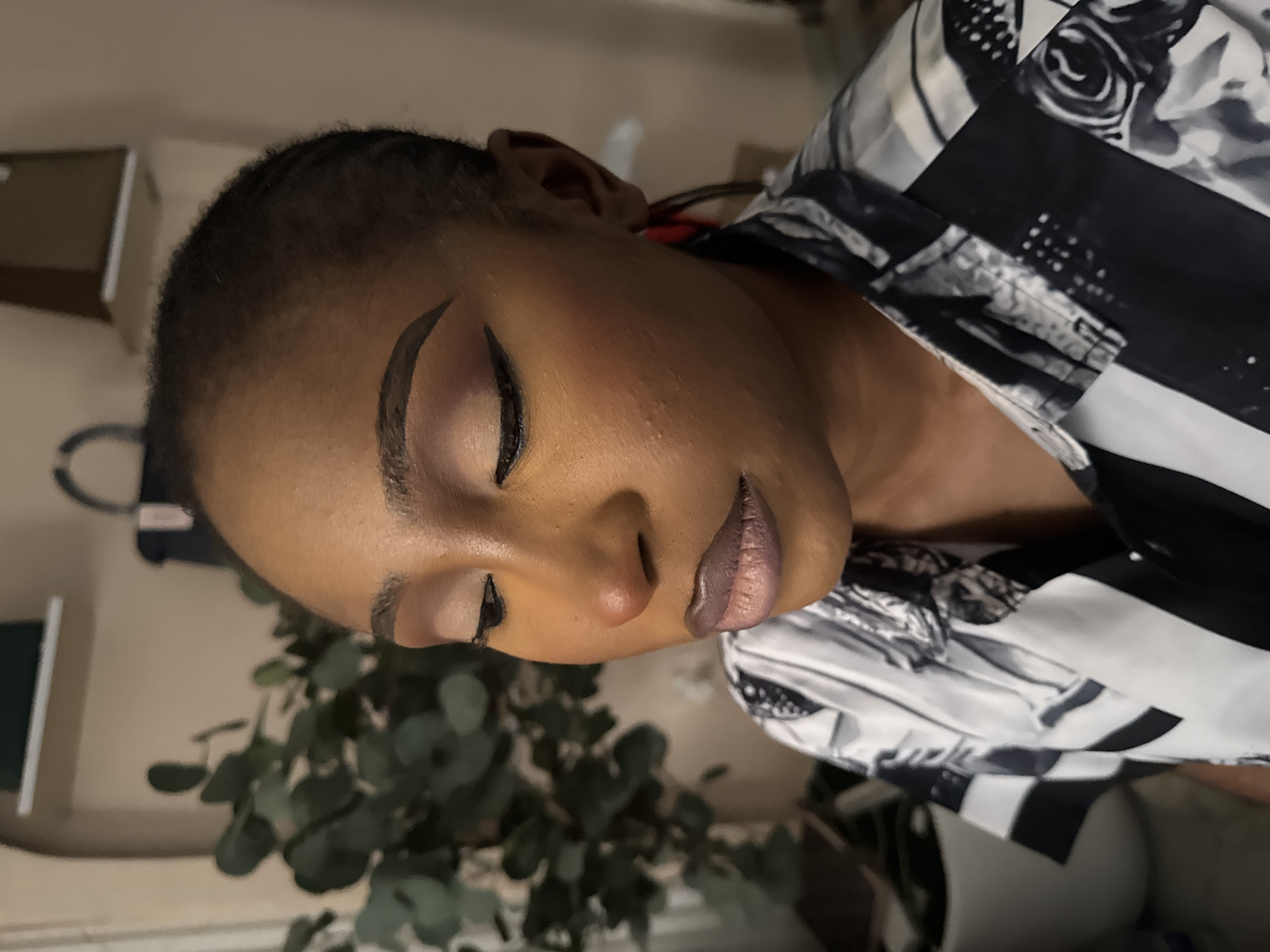 A medium close-up shot shows a young Black woman from the chest up, looking down and to her right with her eyes closed. Her face is angled to the right. She has dark brown braided hair pulled back from her face. Her eyebrows are expertly shaped and filled in. Her eye makeup consists of a light, shimmery shadow on the lid, a darker shadow in the crease, and a winged eyeliner. Her lips are painted with a matte, mauve-toned lipstick, and there is visible texture on her lips. Her skin has a warm undertone with some subtle redness on her cheeks and nose. A few small bumps are visible on her cheek. She is wearing a black and white patterned shirt with a collared neckline. The background is blurred, showing a hint of a plant with large green leaves on the left, and a shelf with an object on it in the upper right corner. The lighting appears to be indoor, coming from the front and slightly to the left, casting soft shadows. There is no indication of the specific location in Abuja, Nigeria.