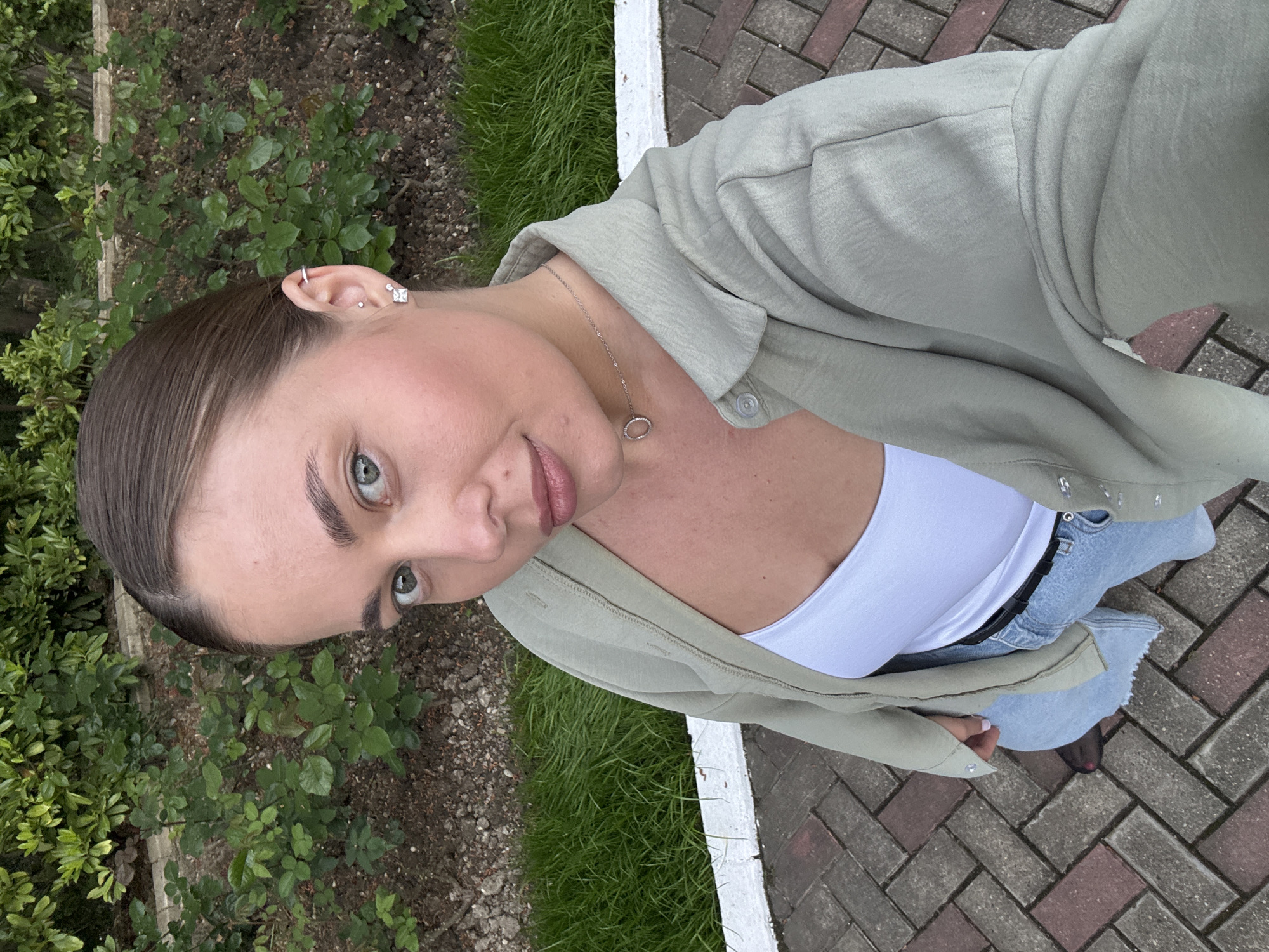 A young woman stands outdoors. She is wearing a light green button-up jacket over a white tank top and light-wash denim jeans with a black belt. She has multiple piercings in her left ear. A silver necklace with a circular pendant hangs around her neck. She stands on a brick pathway, and to her left is a patch of green vegetation. She is likely in a park, and she's taking a selfie.