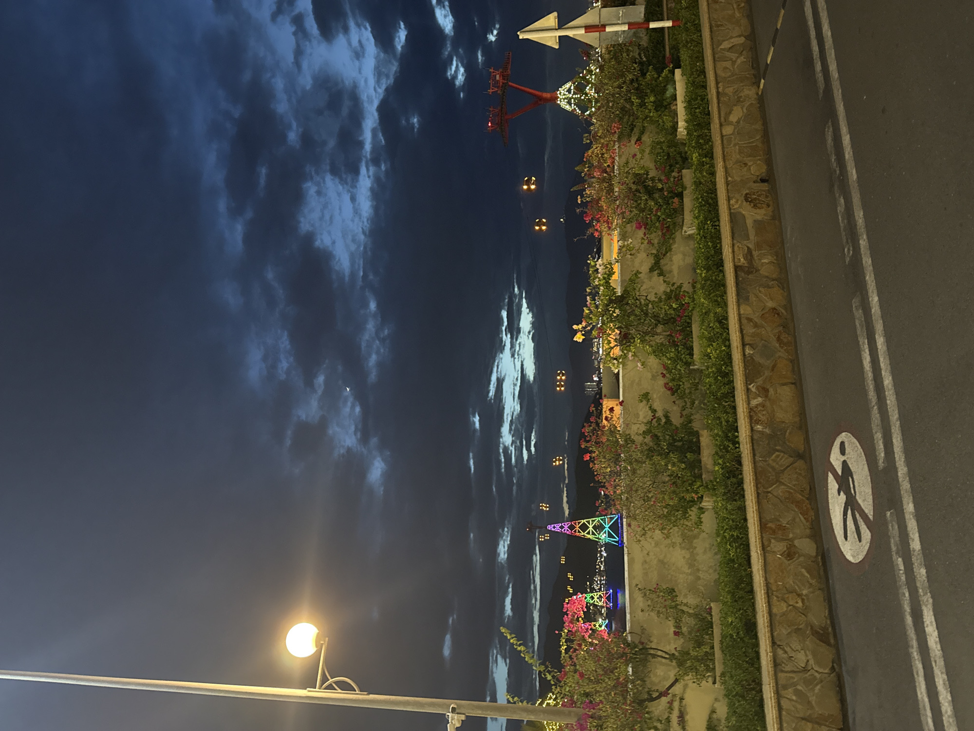 The image depicts a twilight scene in Nha Trang, Vietnam, likely near the cable car station. A street lamp with a lit globe illuminates the foreground left. Beyond it, a low stone wall borders a raised garden bed filled with lush green shrubs and flowering plants in shades of pink and red. In the middle ground, across a body of water, are illuminated structures including two tall, conical towers wrapped in rainbow-colored lights, and a red cable car support tower adorned with white lights. The sky above is a deep blue, filled with dramatic, dark clouds. In the foreground, on the asphalt road, there is a white circular sign with a red border depicting a pedestrian silhouette with a diagonal line through it, indicating "no pedestrians." The scene is lit by artificial lights and the fading natural light of dusk.