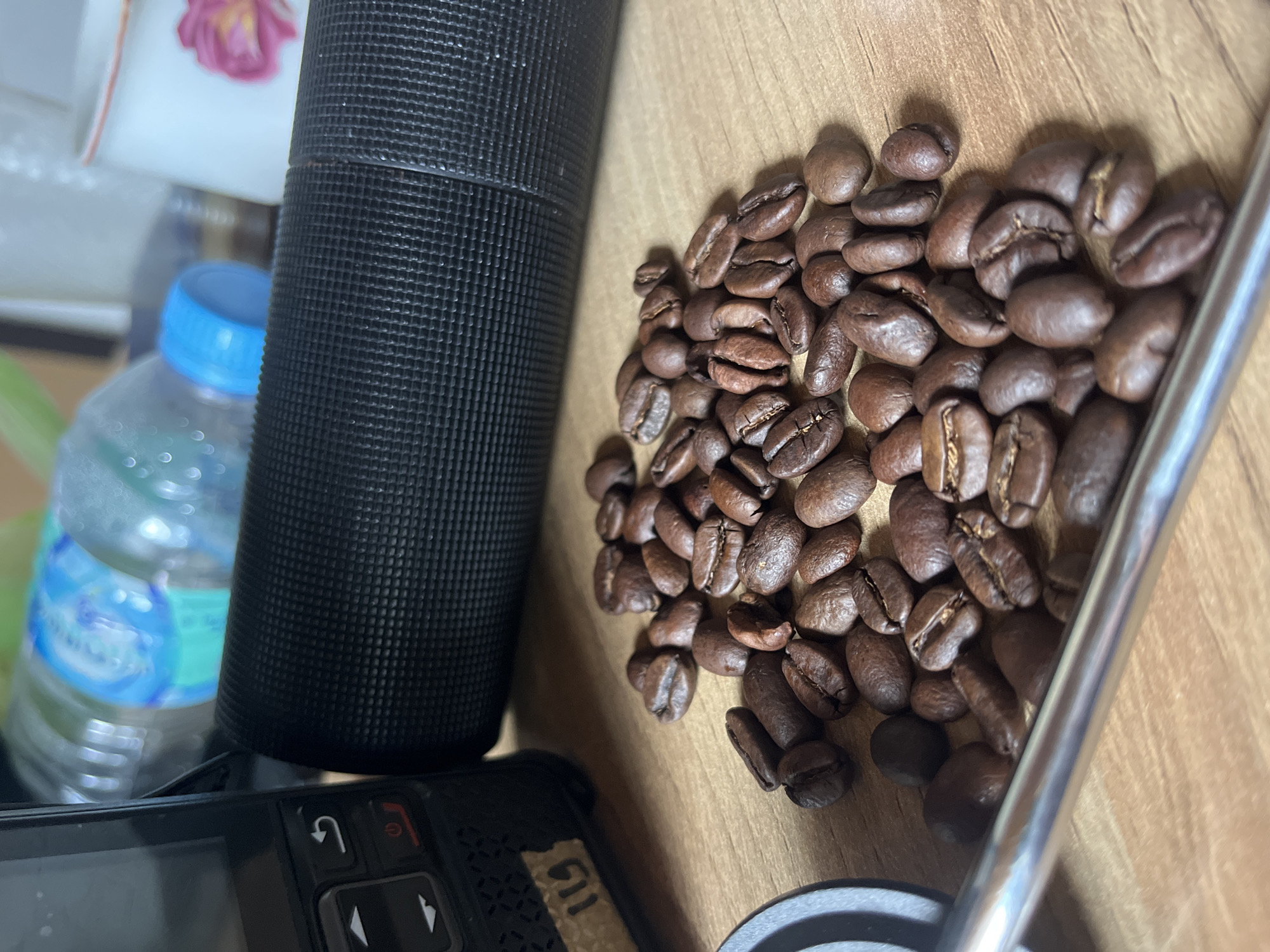 The image shows a close-up view of a wooden surface with several objects placed on it. A pile of roasted coffee beans is located towards the right side of the frame. A black cylindrical object with a textured surface is positioned to the left of the coffee beans. Behind the cylindrical object is a clear plastic water bottle with a blue cap. In the bottom left corner, a black electronic device with buttons and a screen is partially visible. The environment appears to be indoors. Location information (Tambonbaroh, Indonesia) is explicitly provided.