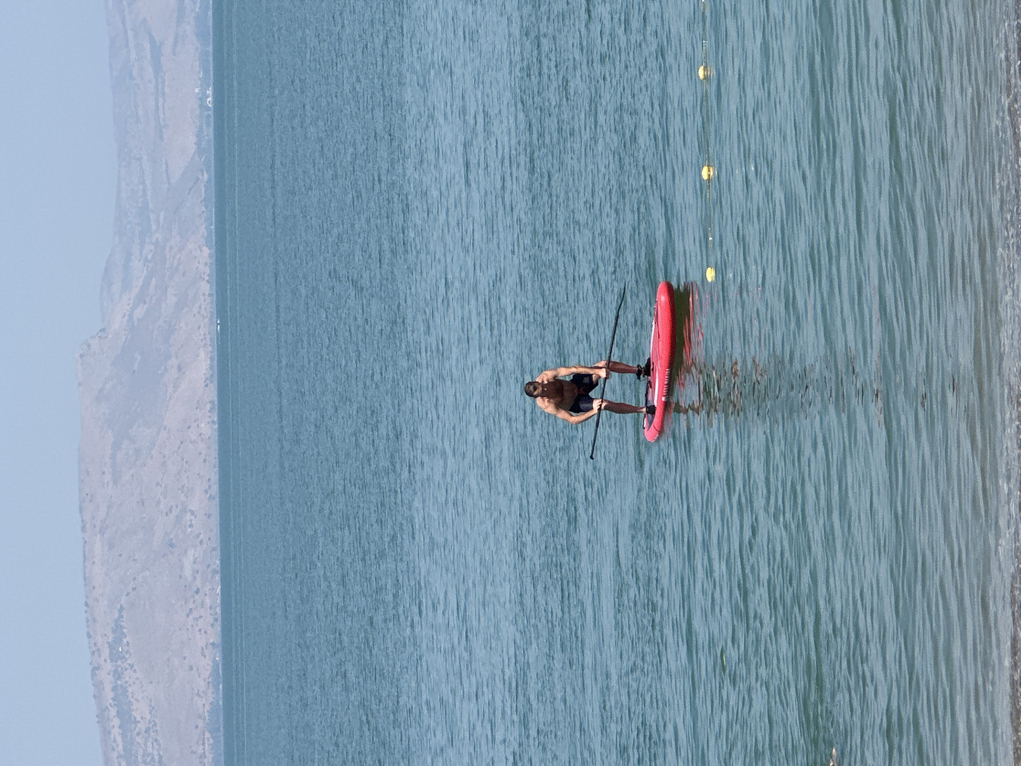 A man is paddleboarding on a large body of water with a shoreline in the foreground and a mountain range in the background. The man is wearing dark shorts and is shirtless, with his hair light brown. He is standing on a red paddleboard and holding a paddle. The water is calm with slight ripples, and the sky is clear and light blue. The mountain range in the background appears arid with sparse vegetation. The shoreline is visible at the bottom of the image, showing sand and pebbles. There are several yellow buoys with a rope connecting them visible in the water. This scene is likely at the Sea of Galilee in the Emek HaYarden Regional Council, Israel, given the distinctive landscape.