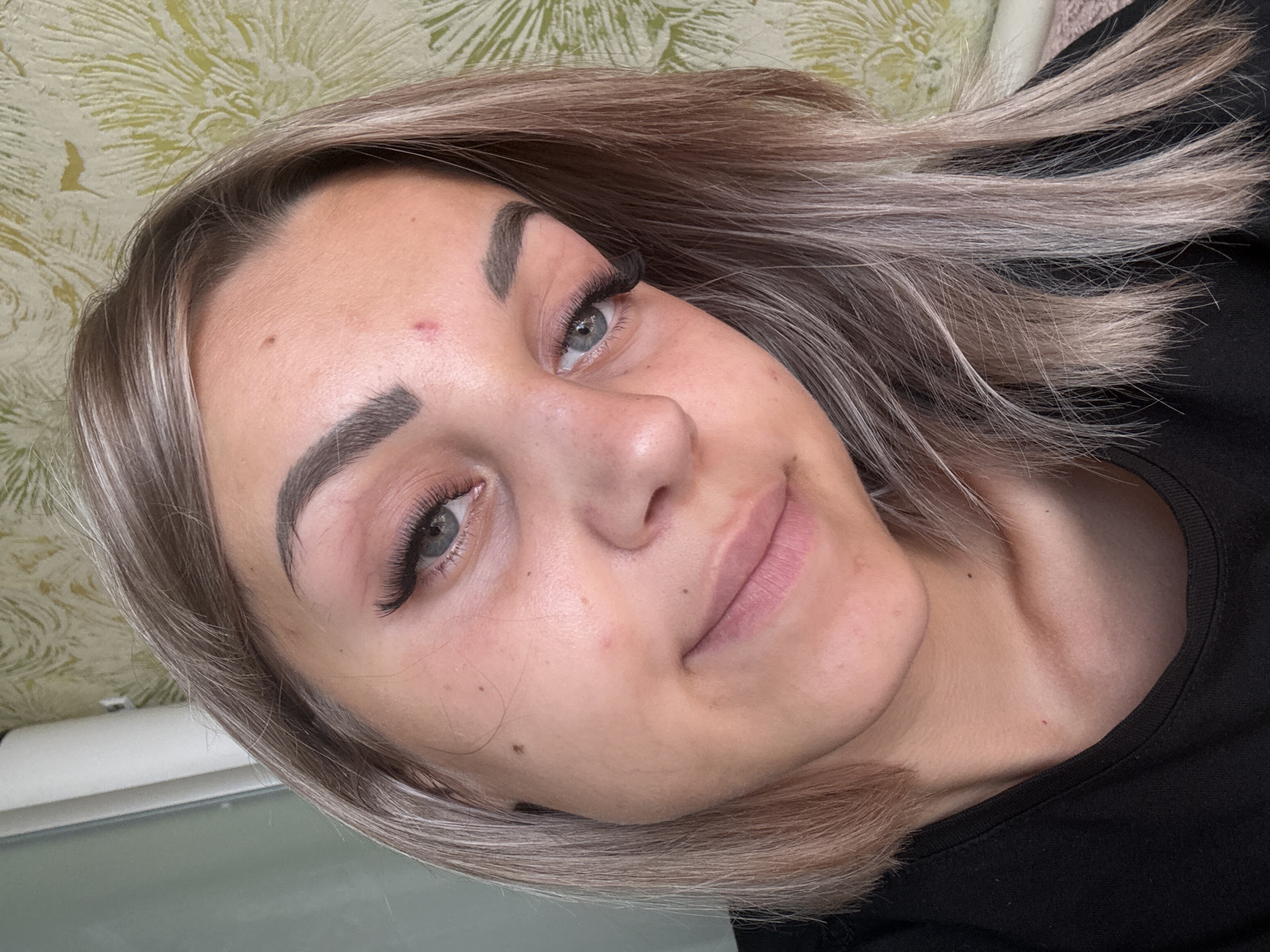 A close-up, selfie-style portrait of a young woman is captured in an indoor setting. The woman has light brown, layered hair with lighter streaks, which frames her face. She is looking directly at the camera with a slight smile. Her eyes are blue, and she has prominent false eyelashes. Her eyebrows are dark and well-defined. Her skin appears fair with some visible pores and a few minor blemishes, including a small red spot on her forehead and a few darker moles on her face. She is wearing a black t-shirt, and the collar is visible.
The background is out of focus but appears to be a wall with a light green, floral-patterned wallpaper. To the left of the woman's head, a portion of what looks like a white door or window frame is visible. The lighting suggests it is daytime. The image does not contain any overt indications of the city of Krasnogorskoye, Russia.