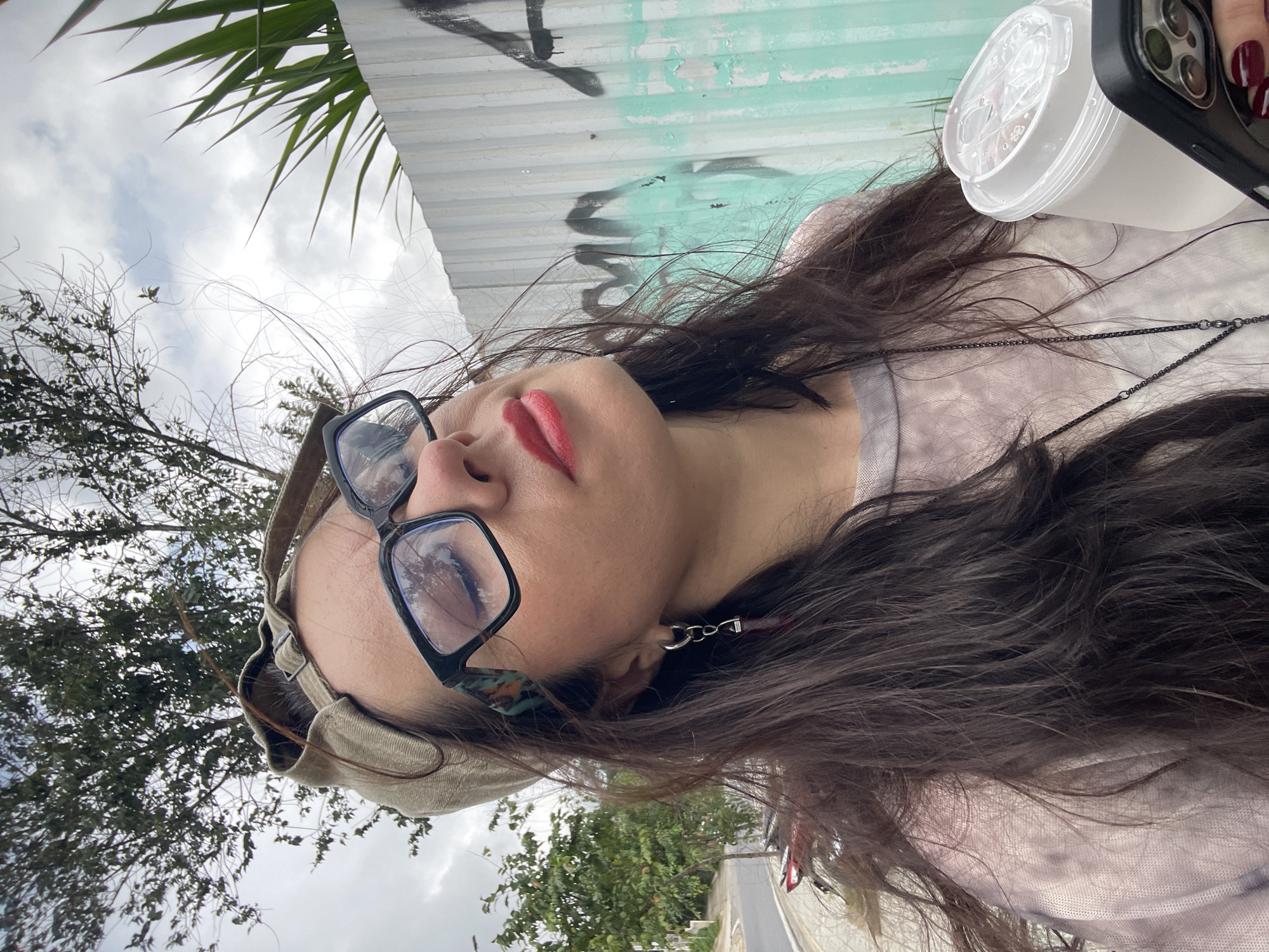 The image is a selfie taken outdoors, possibly in Da Lat, Vietnam, due to the presence of a corrugated metal fence, palm fronds, and a tree with sparse foliage typical of the region. A woman is in the foreground, wearing glasses, a backward-facing baseball cap, and a light-colored t-shirt with a tie-dye pattern. She is holding a clear plastic cup with a white lid in her right hand and a black smartphone in her left. Her eyes are closed, and her lips are painted red. Her dark hair is long and slightly windblown. The background features a cloudy sky, trees, a street, and the aforementioned fence with graffiti. The overall impression is of a casual moment captured outdoors on a slightly overcast day.