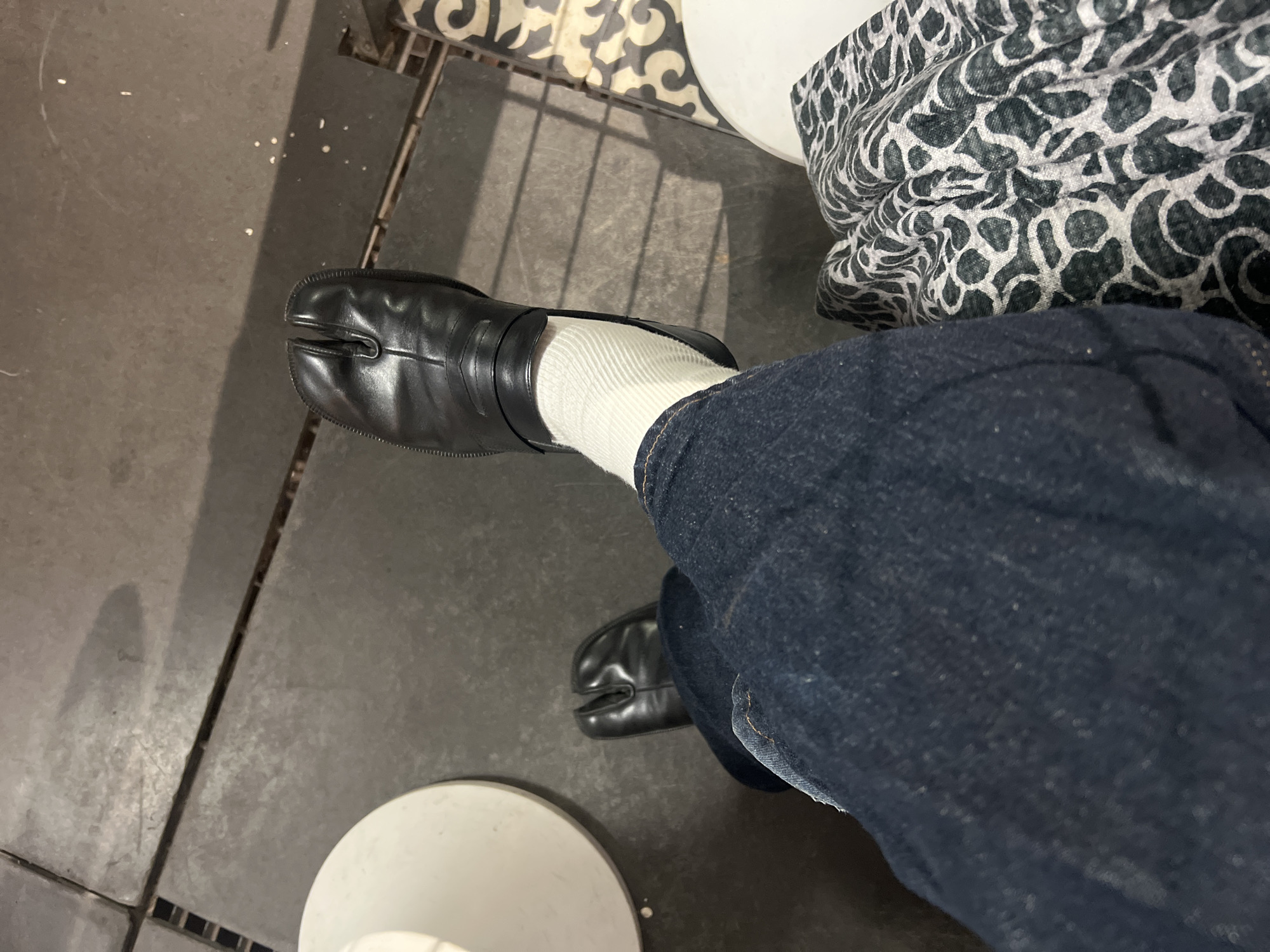 The image captures the lower body of one individual from a top-down perspective, situated in an indoor environment in Tel-Aviv, Israel. The subject is wearing dark blue denim jeans, white ribbed socks, and black leather split-toe shoes (Tabi style). The left leg is extended forward, showcasing the foot with the shoe and sock, while the right leg is bent. A piece of fabric with a black and white or light grey abstract, possibly animal print, pattern is visible on the right side of the frame, partially covering the subject's right leg. The flooring consists primarily of large, dark grey, rectangular tiles with dark grout lines. On the far right, the tiling transitions to a section with decorative black and white patterned tiles. A white circular object, likely the base of a table or stool, is partially visible on the left side of the frame. Small white particulates are scattered on the dark floor tiles in the upper right quadrant. Shadows are cast by the extended leg and shoes onto the tiled floor, suggesting an overhead light source. No specific identifying markers for a precise Tel-Aviv location are discernible from the visual information provided.