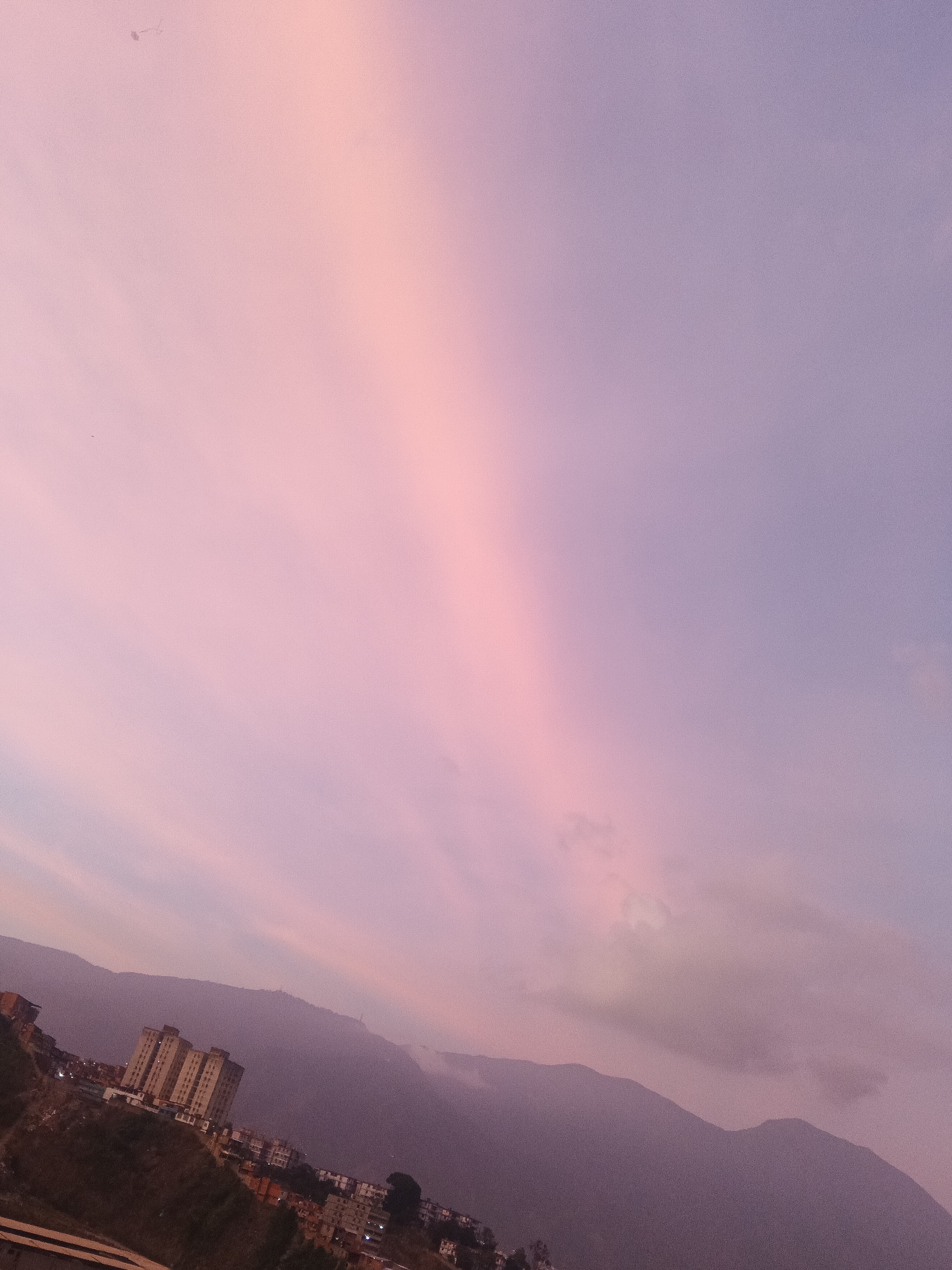 The image depicts a pink and purple sky over Caracas, Venezuela. The foreground features a hillside cityscape with buildings. In the background, there's a mountain range shrouded in mist. The sky shows a prominent vertical band of bright pink light. No people are visible in the image.