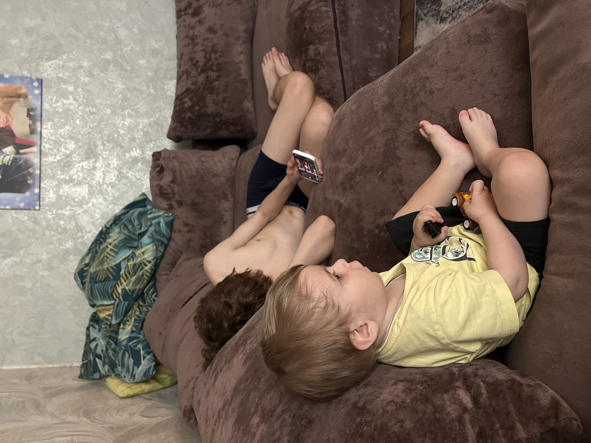 Two children are lying on a brown sofa. One child, wearing blue shorts, is looking at a phone. The other child, wearing a yellow shirt and black shorts, is playing with toy cars. A blanket with a tropical print is next to the sofa. A picture hangs on the wall. The location is Noviy, Russia.