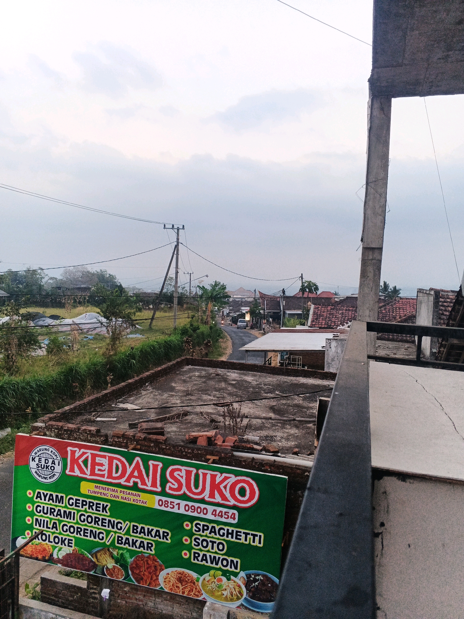 The image captures a view from a balcony or elevated structure overlooking a semi-rural area in Bocek, Indonesia, during what appears to be late afternoon or early morning. In the foreground, a portion of a concrete balcony railing and a pillar are visible on the right. Below the balcony, a rooftop with exposed bricks and debris is present. A large, green banner with red and white text is prominently displayed on the left side, advertising "KEDAI SUKO" which offers various Indonesian dishes such as Ayam Geprek, Gurami Goreng/Bakar, Nila Goreng/Bakar, Koloke, Spaghetti, Soto, and Rawon. The banner also includes images of some of the food items and a phone number: 0851 0900 4454.
Beyond the banner, the scene opens up to a village landscape. A paved road curves through the settlement, with several houses featuring tiled roofs. Some houses have white or light-colored walls, and others have darker brickwork. Trees, including palm trees, are scattered throughout the area. There are utility poles with power lines crisscrossing the sky, indicating the presence of electricity. In the mid-ground, there are fields with some covered structures, possibly agricultural products or temporary shelters. The sky is overcast with soft, diffused light, suggesting a cloudy day. The overall impression is of a quiet, everyday scene in a small Indonesian town. There are no people clearly visible in the frame, and no specific actions or interactions are depicted.
