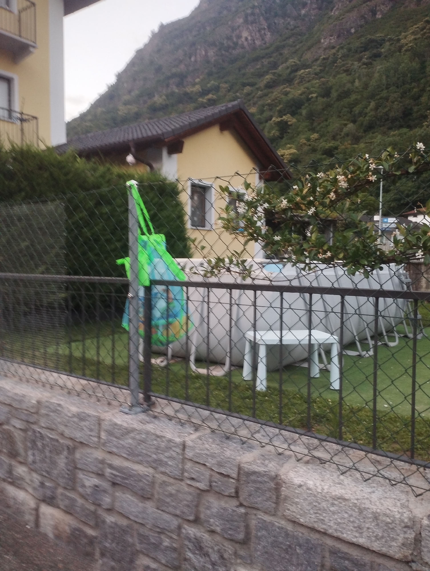 A grey chain-link fence borders a grassy yard in front of a yellow house with a dark-tiled roof, which is nestled against a steep, tree-covered mountainside. A large, above-ground swimming pool is set up in the yard. In the foreground, a wall of rough-hewn grey stones runs horizontally across the image. A green mesh bag hangs from a fence post. White flowers bloom on a bush in front of the pool. The sky is overcast.