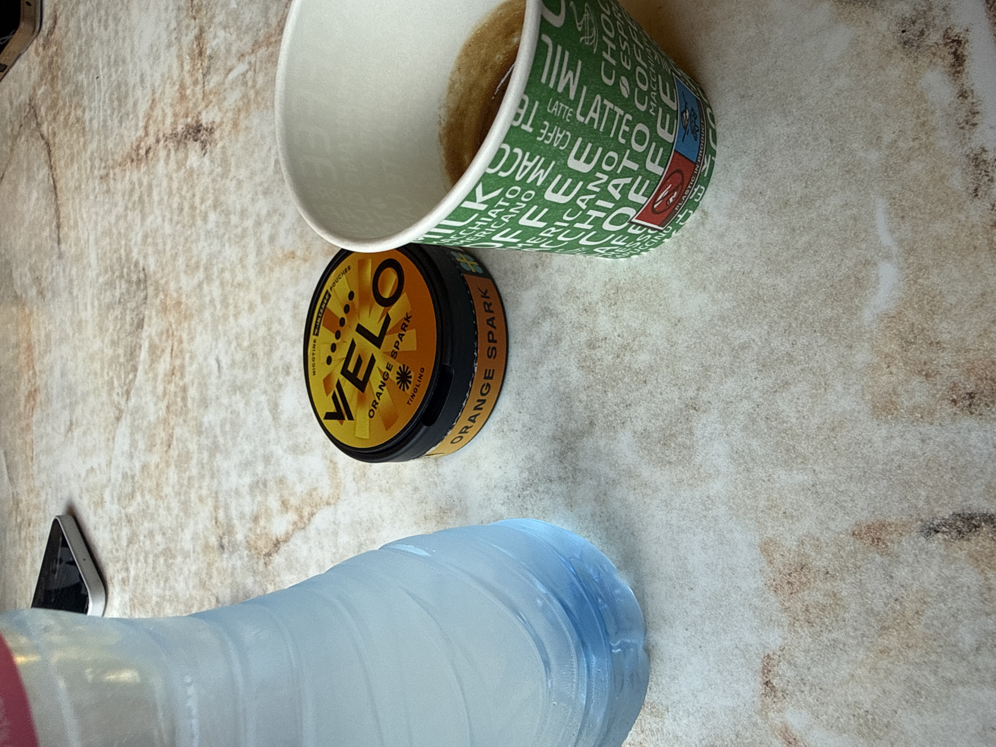 The image displays an overhead shot of a light-colored, mottled tabletop, resembling marble or granite, with three primary objects arranged on its surface. On the left, a partially filled, light blue, ribbed plastic water bottle stands upright. In the center-right, a circular, black and yellow container for VELO nicotine pouches is visible, labeled "ORANGE SPARK" with "NICOTINE INTENSE POUCHES" and "TINGLING" also present. To the far right, a green and white disposable paper coffee cup, containing a liquid with a foamy crema layer, is positioned. The cup's exterior is patterned with repeated text including "MILK," "COFFEE," "LATTE," "CAPPUCCINO," "MACCHIATO," and "AMERICANO." In the upper background, out of focus, the corner of a white-backed smartphone is partially visible. No individuals are present in the scene. Based on the visual evidence within the image, there are no discernible features or specific identifiers to confirm the location as Blagoevgrad, Bulgaria.
