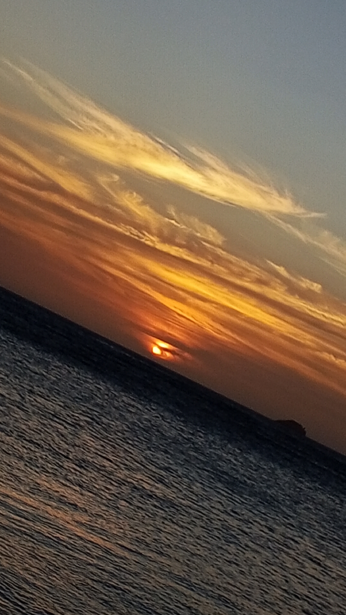 The image shows a sunset over the ocean. The sky is filled with streaks of yellow and orange clouds. The sun is partially obscured by the horizon, creating a warm glow. The water is dark and rippled. In the distance, there is a small island. The scene is angled, suggesting the photo was taken at a tilt. The location is likely Pedro González, Venezuela, given the prompt.