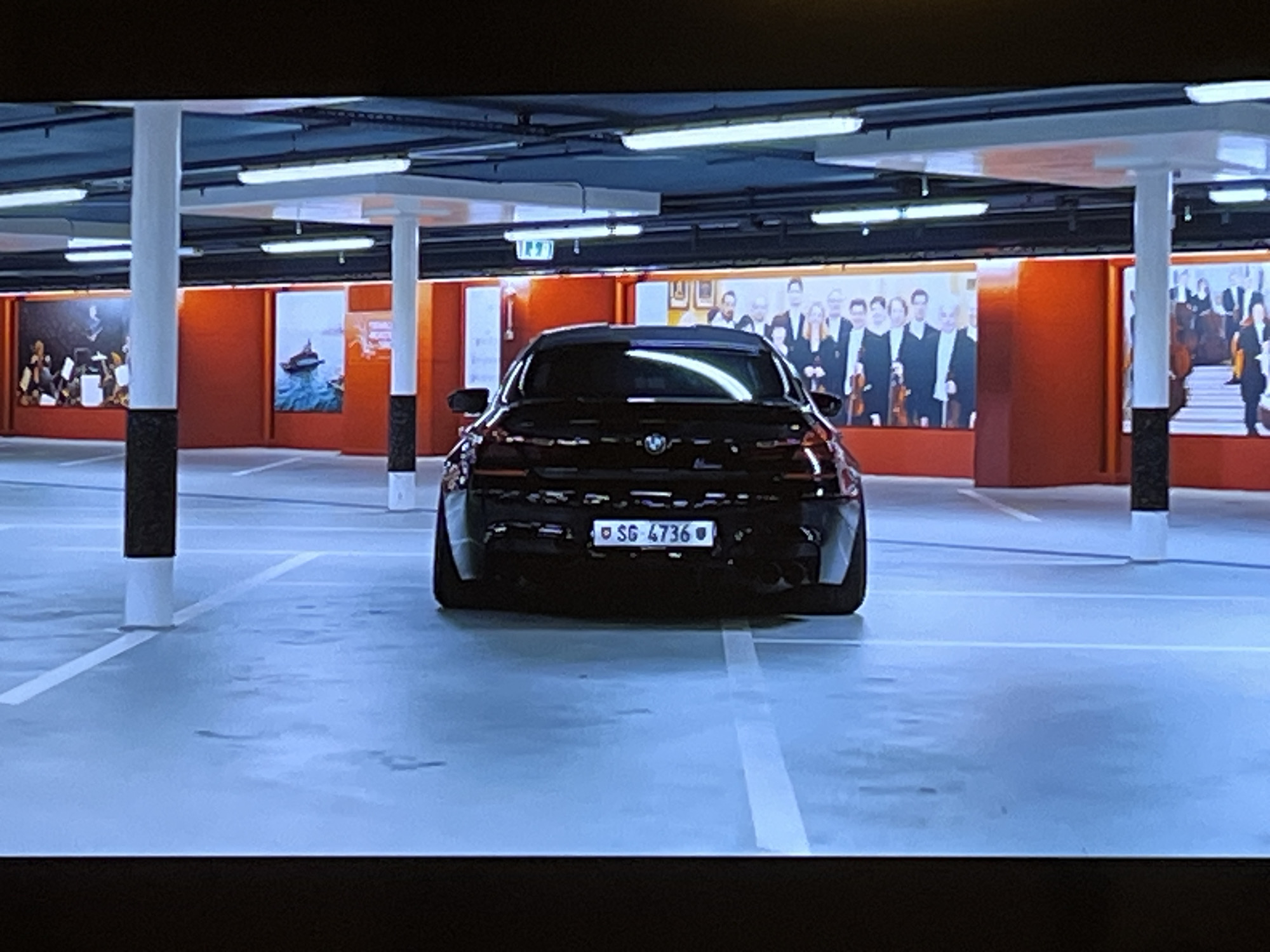 A black BMW is parked in a brightly lit underground parking garage. The car's license plate reads "SG 4736". The garage has white painted lines on the floor, white columns, and fluorescent lighting. The walls in the background feature large murals depicting orchestral musicians and a boat on water. The overall impression is that of a modern and clean indoor parking facility. The location of Kharkiv, Ukraine is not discernible from the image.