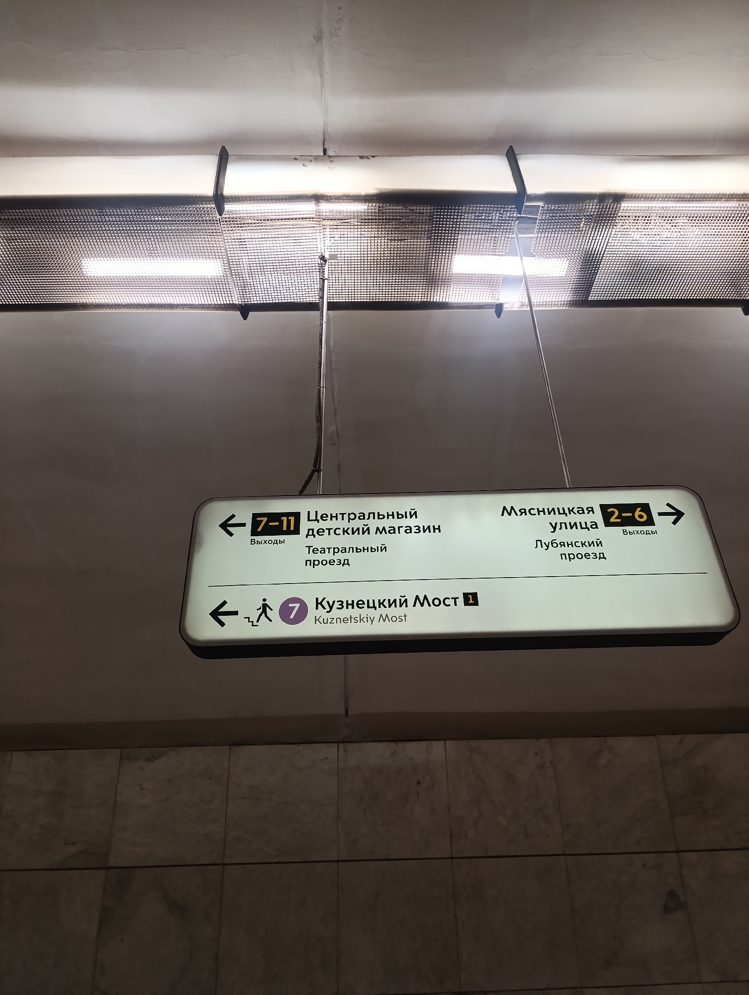 Here is a detailed description of the image:
The image shows a subway station sign in a city where Cyrillic script is used, likely in Russia. The sign is rectangular, white, and illuminated from within. It is suspended from the ceiling by metal rods. The sign indicates directions to various destinations. To the left, it lists "Central Children's Store," "Teatralny Passage," and the "Kuznetsky Most" (Kuznetsky Bridge) metro station with line number 7. To the right, it points to "Myasnitskaya Street" and "Lubyansky Passage." The sign also denotes exits numbered 7-11 on the left and 2-6 on the right. The background features a ceiling with lighting fixtures and a tiled wall below the sign.