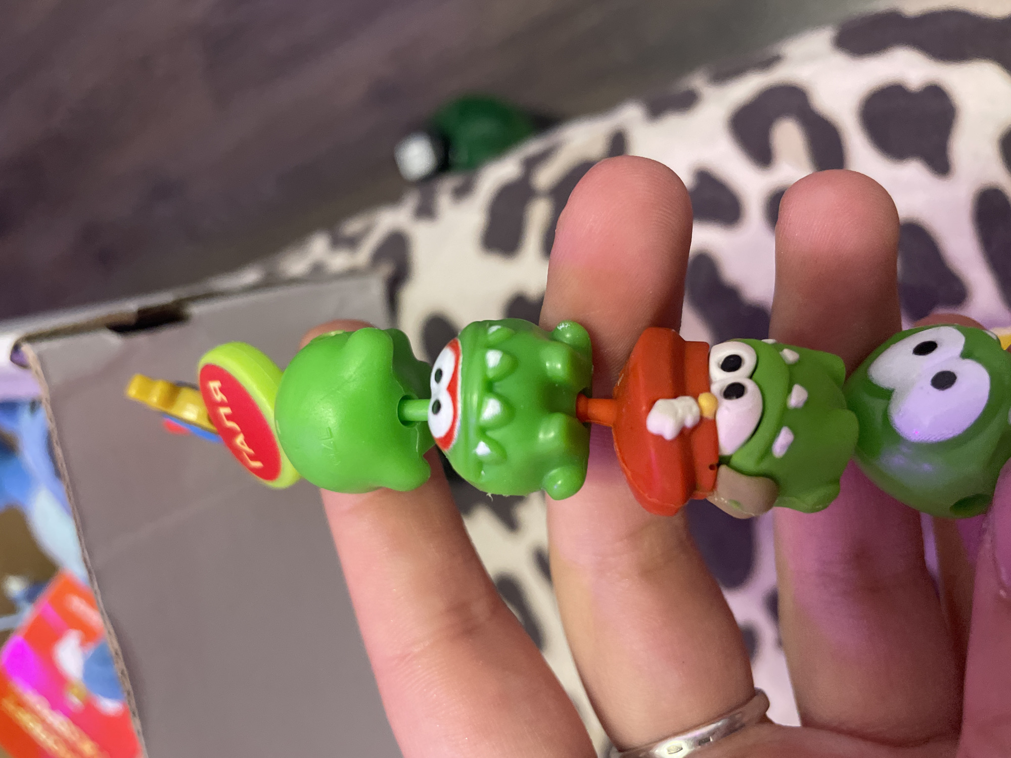A close-up, eye-level shot shows a person's hand holding a string of green, cartoonish figurines, likely part of a keychain or toy. The figurines are stacked vertically, with each figure having a distinct facial expression and some wearing accessories like hats. The hand is lightly touching the figurines, and a silver ring is visible on one of the fingers. In the blurred background, a cardboard box with colorful elements and a patterned fabric are discernible. The scene appears to be indoors, with no indication of the city of Kokkorevo, Russia.