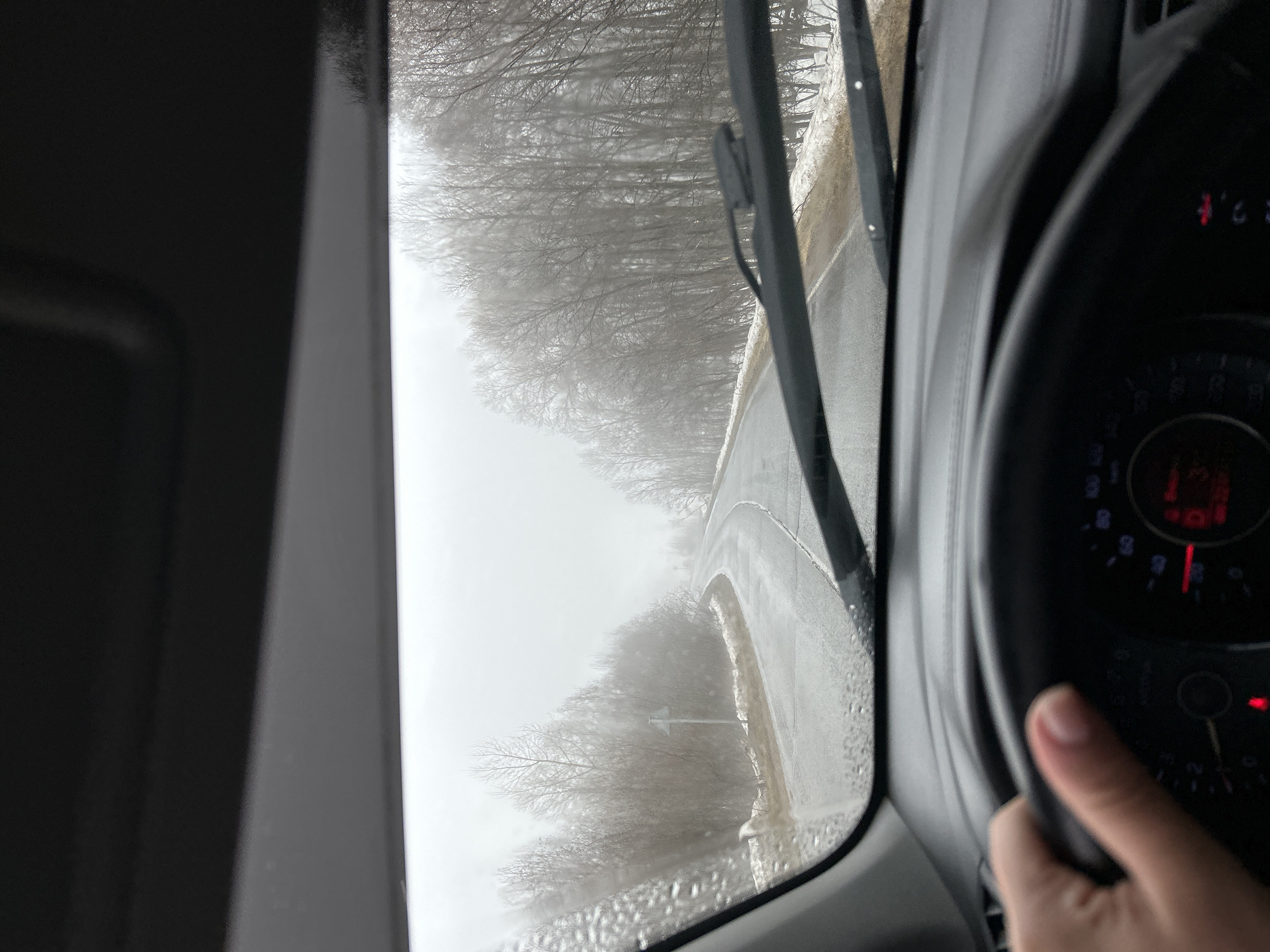 The image shows a view from inside a car driving on a road in Уральское, Russia. The weather is snowy and visibility is low. The road is wet and winding, with snow on the sides. Bare trees line the road. The car's windshield wipers are in use. A hand is visible on the steering wheel, and part of the car's dashboard is visible to the right.