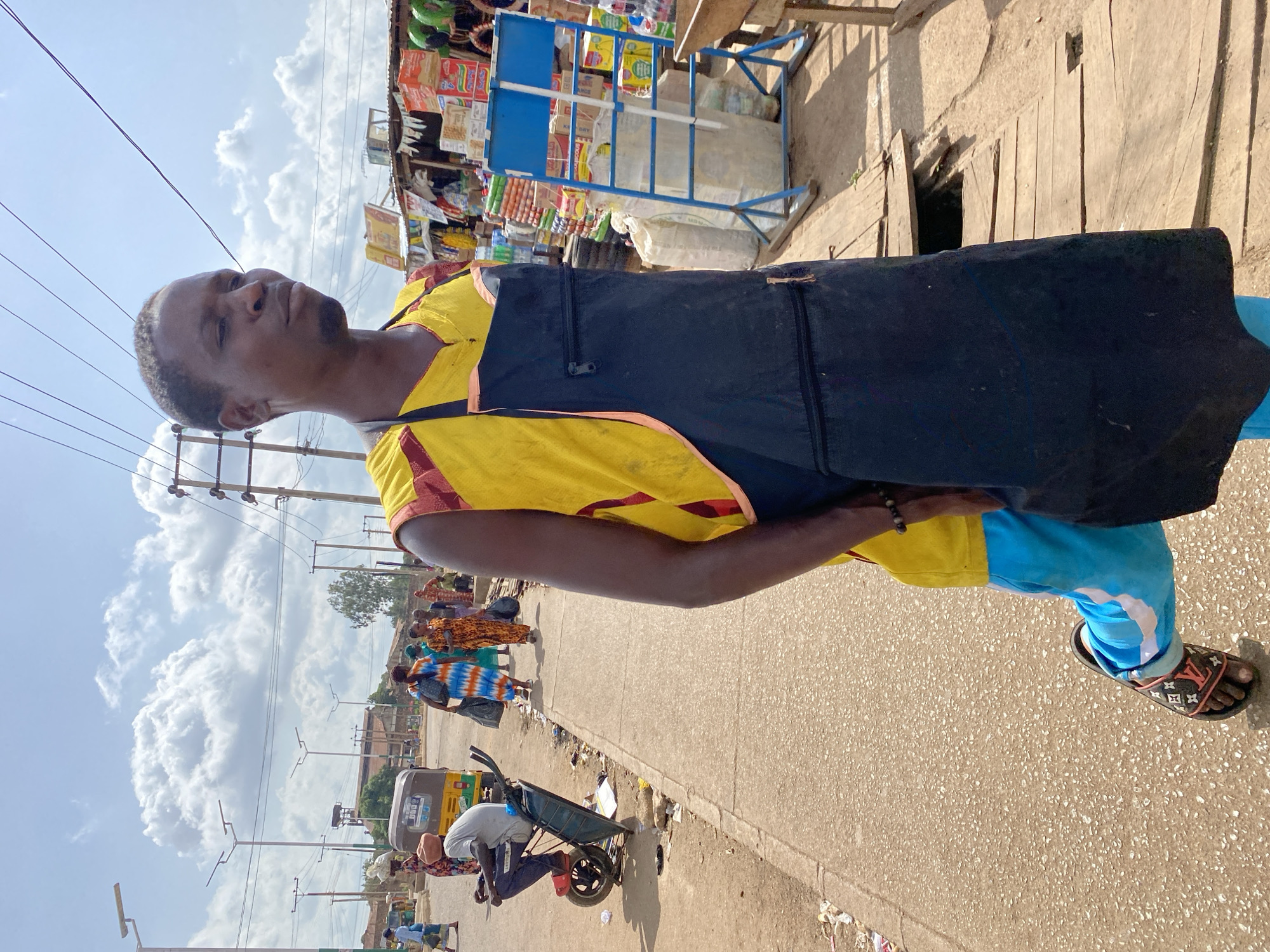 The image depicts a street scene in Bukuru, Nigeria, under a bright, cloudy sky. The foreground is dominated by a man wearing a yellow sleeveless shirt with red trim, a black apron, and light blue pants with white trim. He is wearing distinctive black sandals with an orange and black pattern. His left leg is bent at the knee, and he appears to be in motion.
In the background, several other people are visible. To the left, a man sits on a wheelbarrow that has been modified into a form of transportation, with a motor attached to one wheel. He is wearing a beige turban and dark pants. Further down the road, a group of people, mostly women, are walking. One woman wears a vibrant blue and white tie-dye dress, another a patterned orange dress, and another a patterned pink and white dress. They are carrying bags.
The environment is a market area. To the right of the frame, stalls are set up with various goods, including stacked boxes of what appear to be food products and bags of goods on display. A blue metal stand holding more items is also visible. The road surface is asphalt, and a concrete sidewalk runs alongside it. Debris and litter are present along the edge of the road and sidewalk. Overhead, power lines and streetlights are evident. The overall impression is of a bustling local market and street scene in Nigeria.