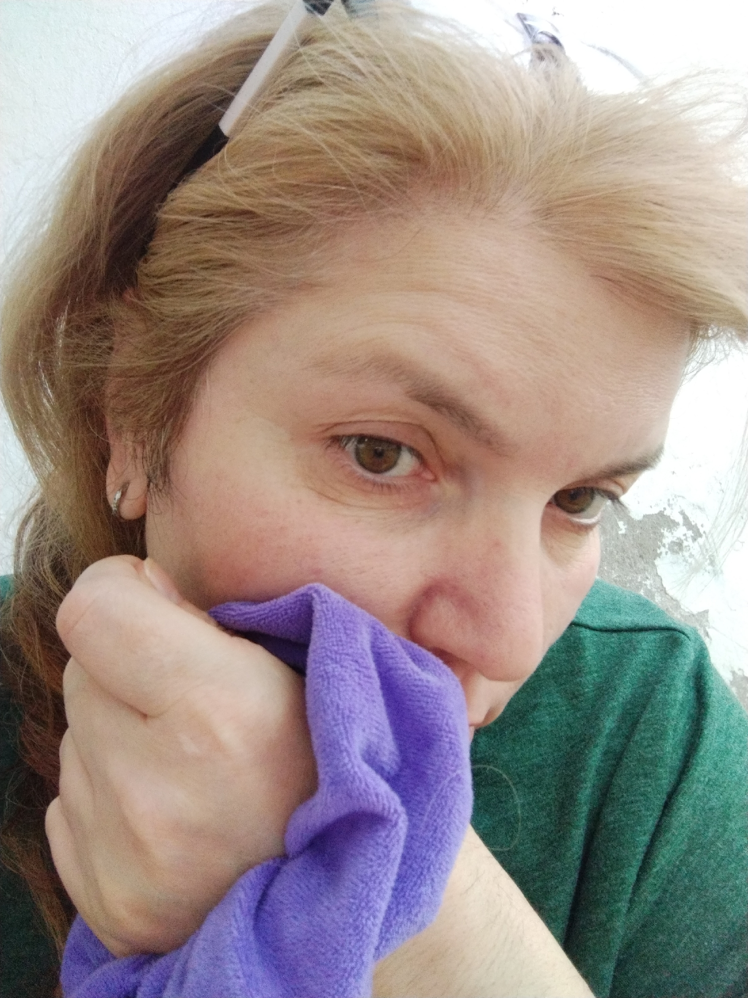 A fair-skinned woman with light brown hair is partially obscuring her face with a small, purple towel. She is wearing a green t-shirt and has an earring in her left ear. A hair clip is visible in her hair. The background appears to be an interior wall with peeling paint. The exact location within Al Mas'oudi, United Arab Emirates, cannot be determined from the image.