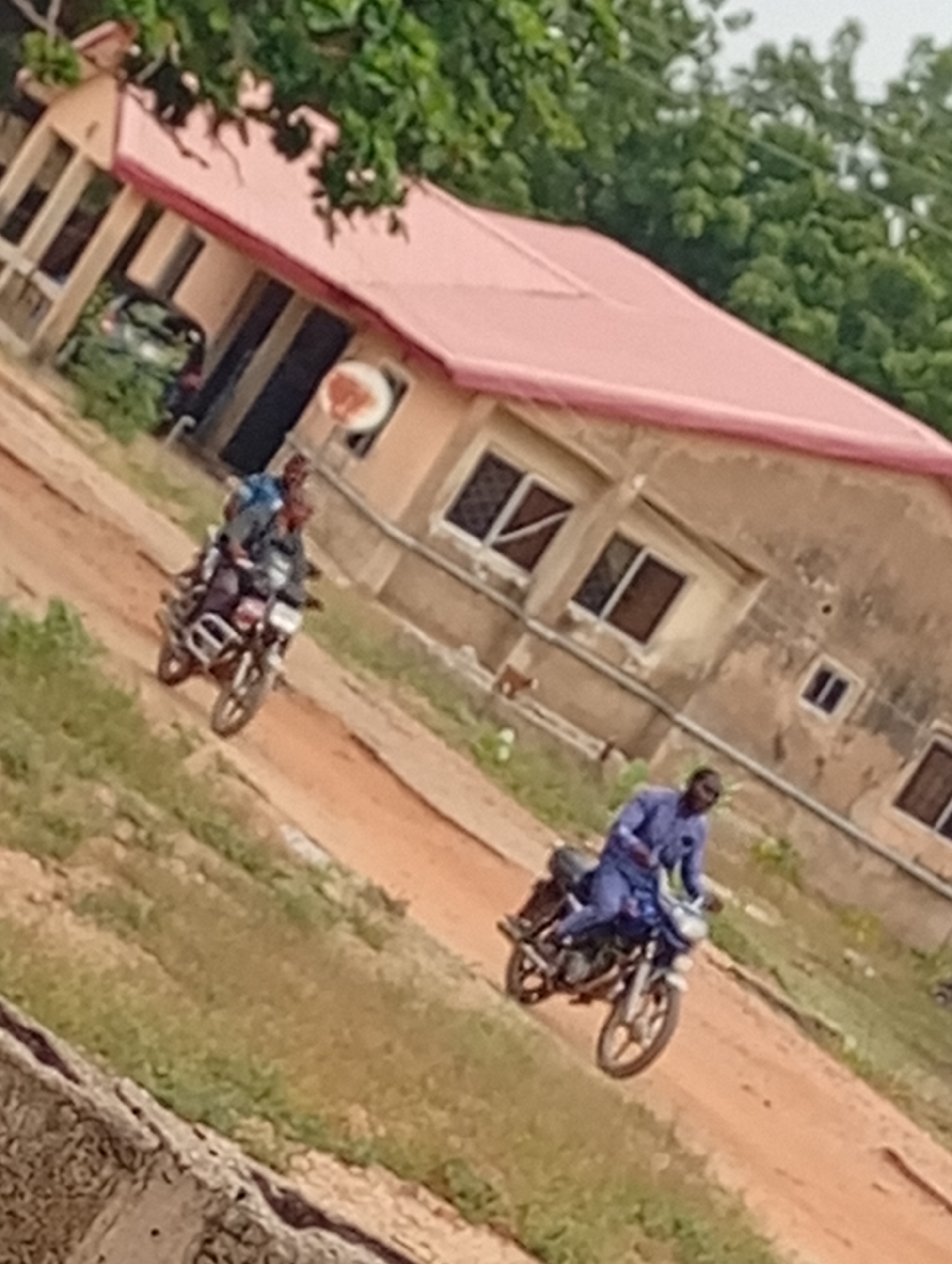 The image depicts two motorcycles on a dirt road in front of a building with a pink roof. On the first motorcycle, two people are riding, while on the second, a single person is riding. The location appears to be an urban setting in the city of Unknown, Unknown. The building has several windows and what looks like a satellite dish attached. Trees and greenery are visible in the background and around the building.