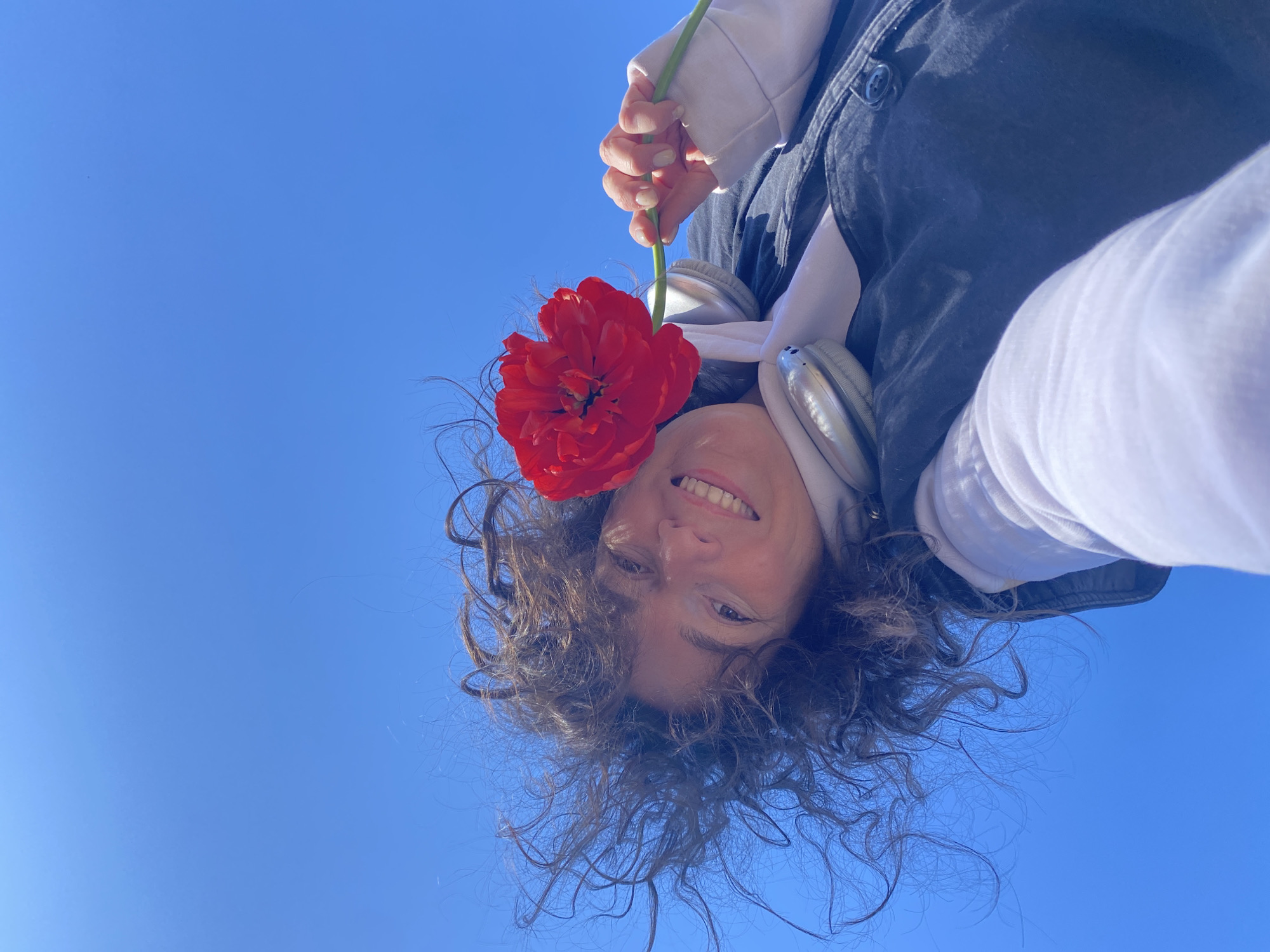 A person with curly brown hair is holding a red flower. The person is wearing a white shirt and a black jacket and headphones around their neck. The background is a clear blue sky. The photo appears to be taken outdoors, looking upwards. The location is Ольховка, Russia.