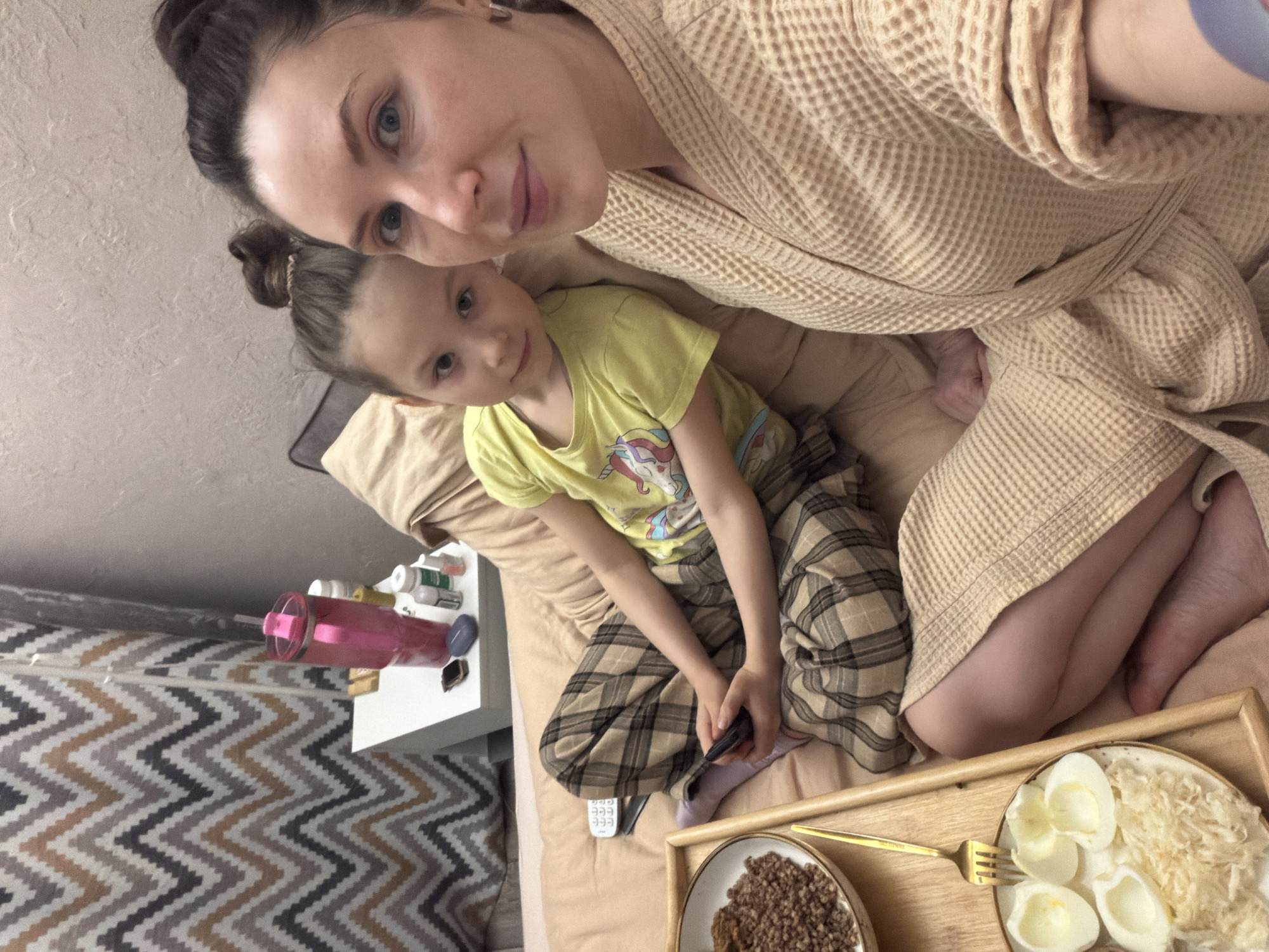 A selfie shows a woman and a young girl in a domestic setting. The woman, wearing a light brown waffle-textured bathrobe, has her dark hair in a bun. The girl is wearing a yellow t-shirt with a unicorn graphic and plaid pajama pants, and she holds a remote control. They are situated on a bed with light brown sheets, and a food tray with buckwheat and sliced eggs is in the foreground. A small white side table with a pink water bottle and various pill bottles sits adjacent to the bed. The background includes a wall with a patterned zig-zag design. The location is likely indoors, possibly in a residential area in Perm, Russia. The woman is taking a selfie.