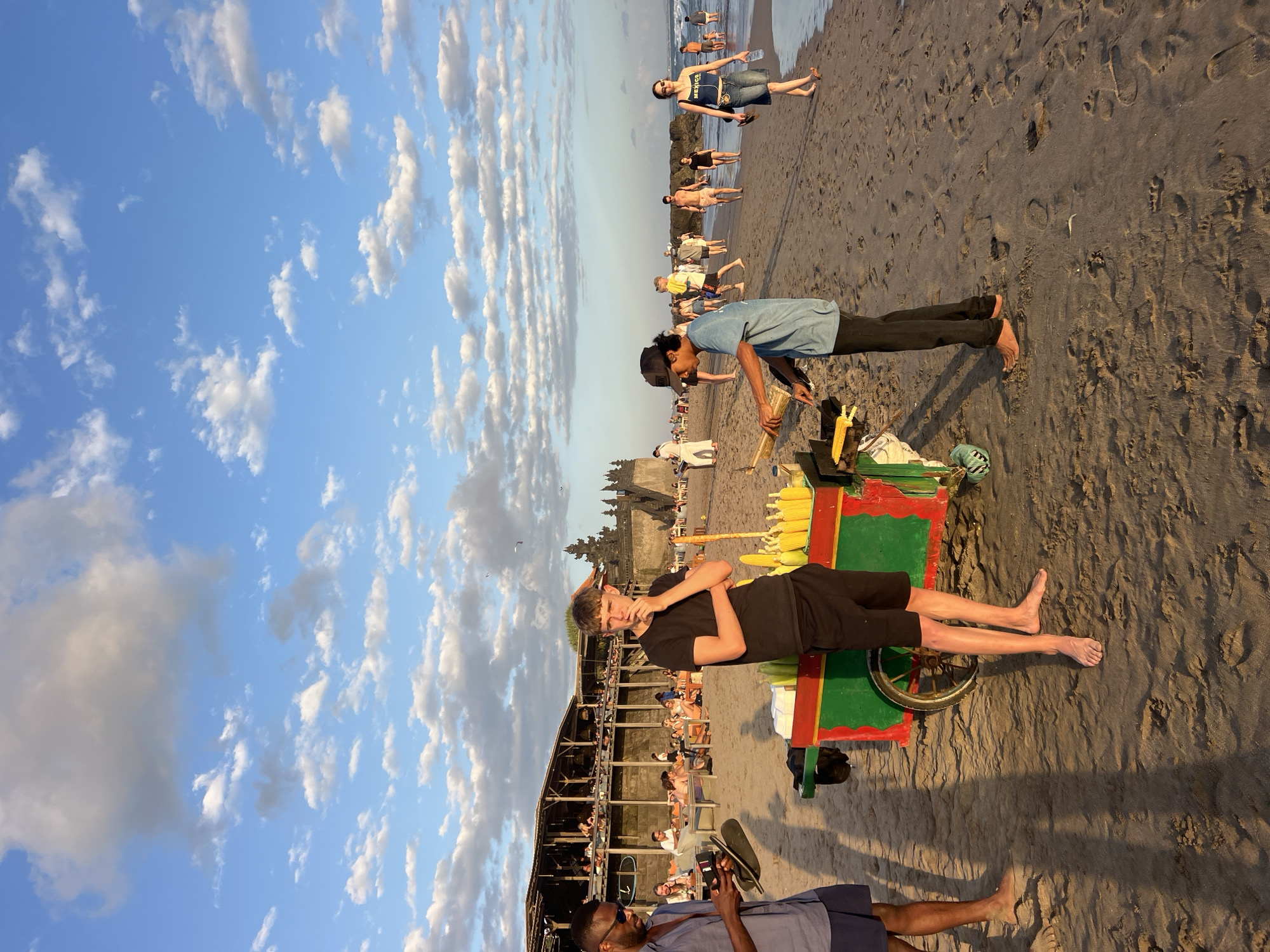 An outdoor scene on a beach in North Kuta, Indonesia, captures a sunset with a vibrant sky filled with white clouds. In the foreground, a colorful food cart with green, red, and yellow accents is positioned on the sandy shore. Two young men stand near the cart. One, wearing a black t-shirt and shorts, stands with his arms crossed and hand on his chin, looking thoughtfully towards the right. The other, in a blue t-shirt and dark pants, is engaged in preparing food at the cart. Further back, a group of people are seen dining at a beachside restaurant with an open-air structure and thatched roof. Additional beachgoers are scattered along the shoreline, with some walking and others relaxing. In the distance, a traditional Balinese temple structure is visible against the horizon, silhouetted by the warm glow of the setting sun. The waves of the ocean gently lap the shore.