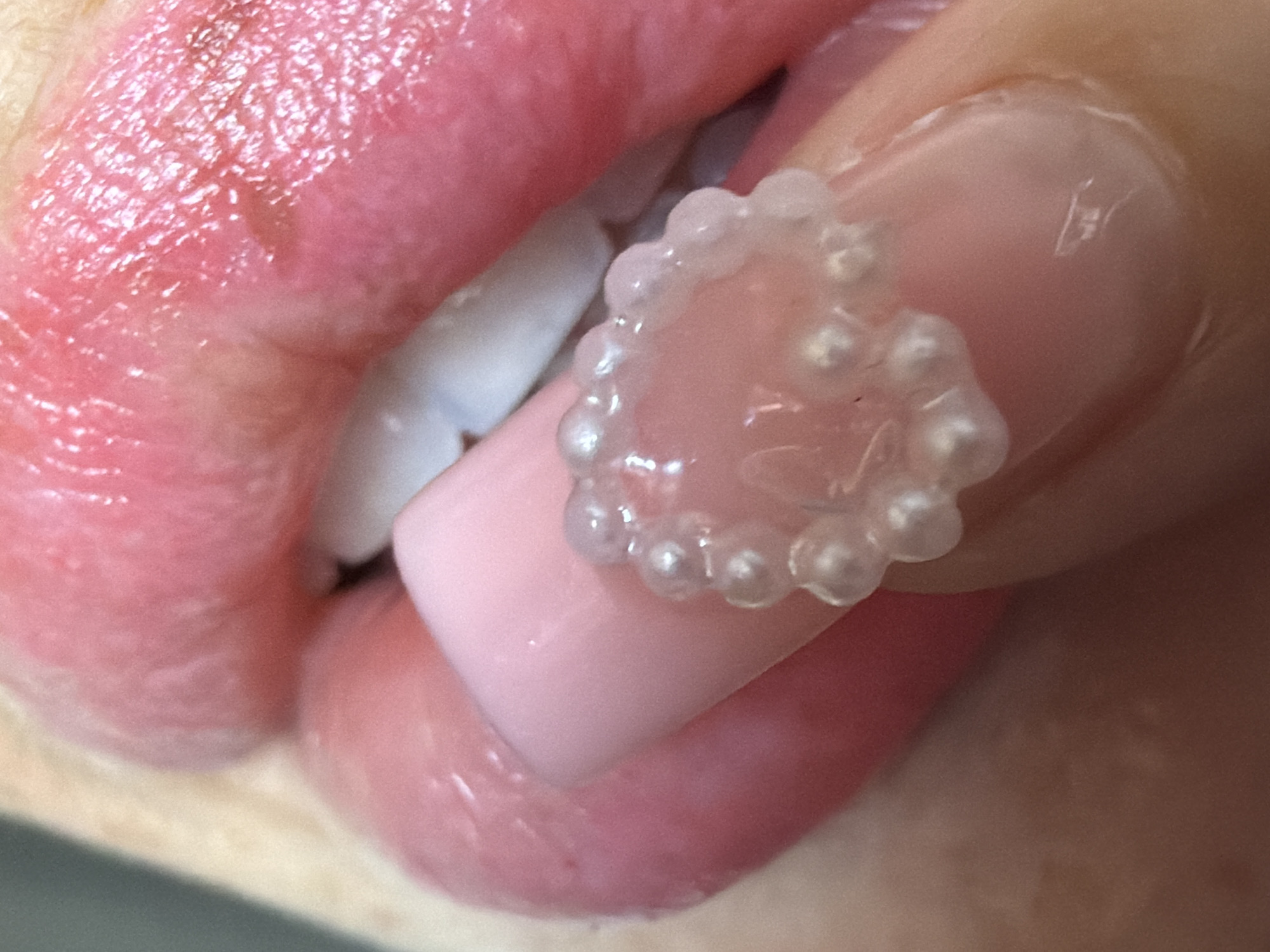 The image is a close-up, overhead view of a person's lips and fingernail. The lips are pink and appear to be slightly chapped or dry. The fingernail is light pink and adorned with a decorative element that resembles a string of small, translucent pearls. The pearl decoration is adhered to the nail, forming a circular shape. The background is blurred and out of focus, suggesting a shallow depth of field. There is no discernible city or location information present in the image.