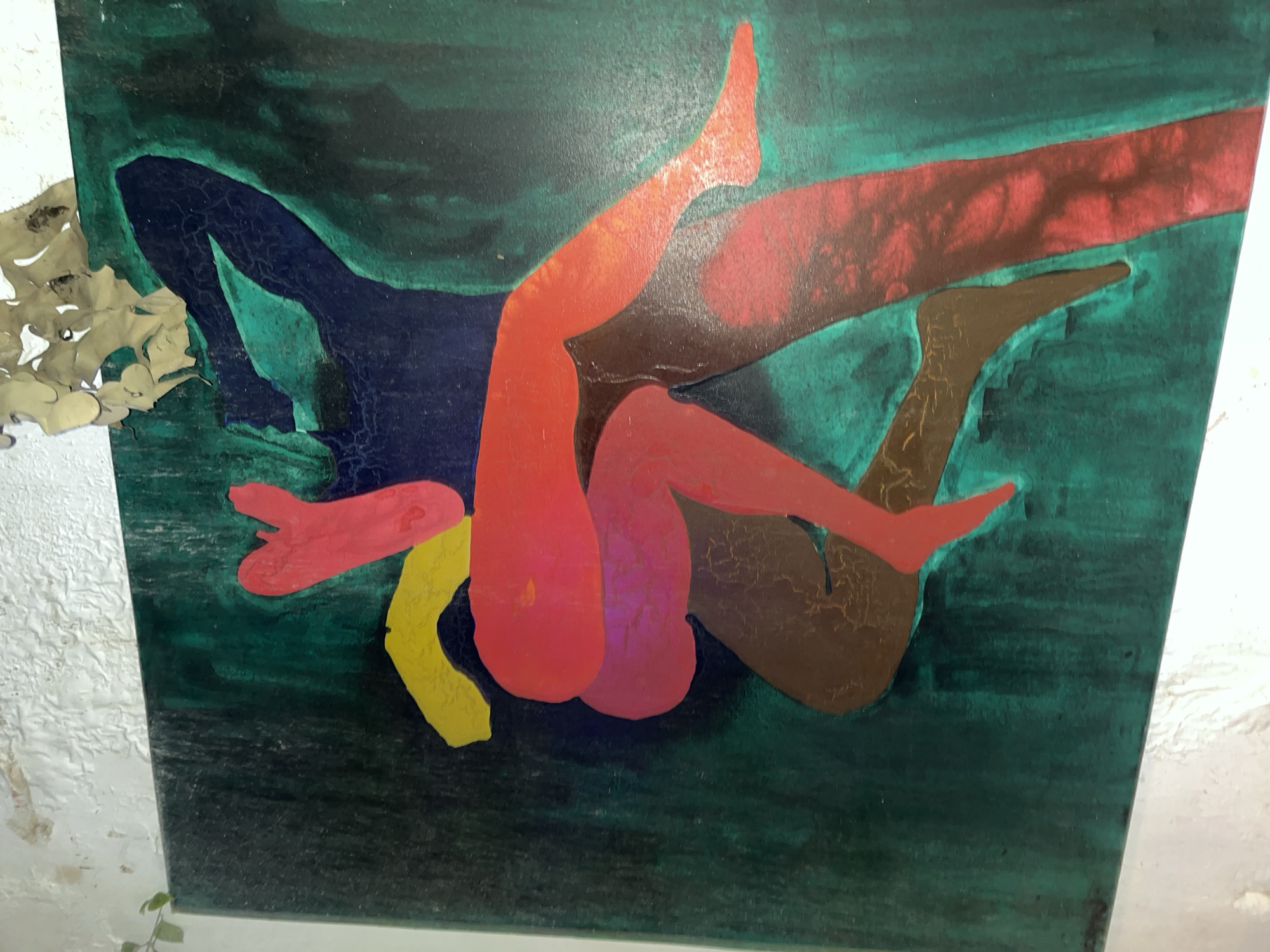 This is a painting depicting two figures in an embrace against a dark green background. The figures are rendered in bold, abstract shapes and colors. One figure appears to be primarily in shades of navy and red, with a section of yellow. The other figure is largely in shades of red and brown. The background has a textured, cracked appearance. Above the painting, a lighter-colored, irregularly shaped object made of what appears to be layered material is attached to the wall. A small green vine with leaves hangs from the top left edge of the frame, near the wall. The painting is affixed to a textured, off-white wall that shows signs of peeling paint. The image appears to be taken from a slightly elevated angle, looking down and to the right. The specific location in Accra, Ghana is not discernible from the image itself.