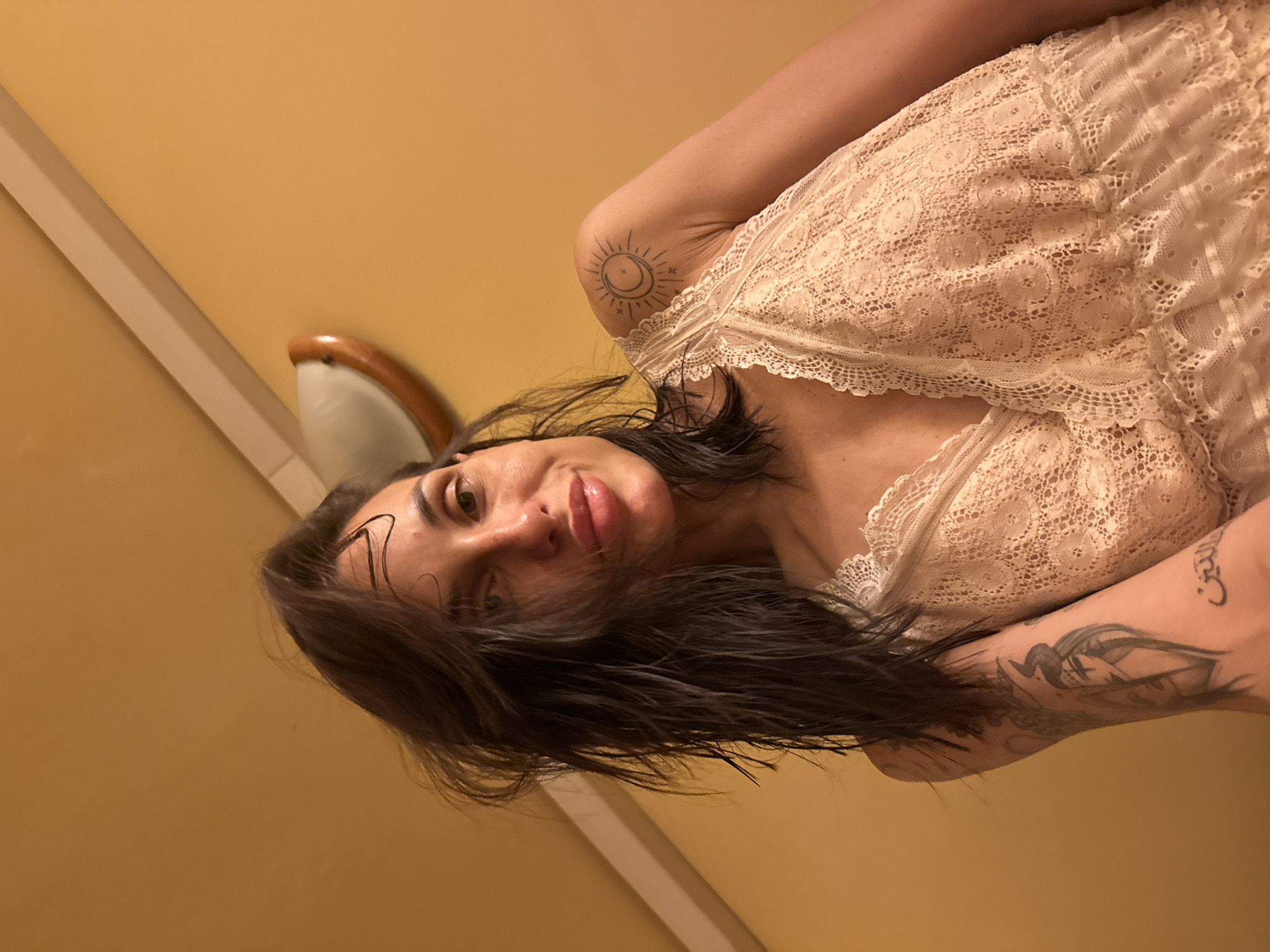 A female subject, appearing to be an adult, is depicted in an indoor setting in the city of Pleven, Bulgaria. She has dark, damp hair that is disheveled, suggesting recent wetting. The subject is looking directly at the camera with a slight smirk or smile. Her lips appear full and glossy. She is wearing a light-colored, possibly beige or off-white, lace camisole with a V-neckline and intricate floral lace patterns. Tattoos are visible on both arms: a minimalist sun and crescent moon design on her right shoulder, and a larger, more detailed tattoo on her left forearm depicting a horned female figure above Arabic script that translates to "the dragon." The background consists of a solid, warm yellow-orange colored wall. A white ceiling beam is diagonally positioned in the upper left, and a wall-mounted light fixture with a curved, frosted shade and a wooden base is visible in the upper right. The image is taken from a low angle, looking upward at the subject.