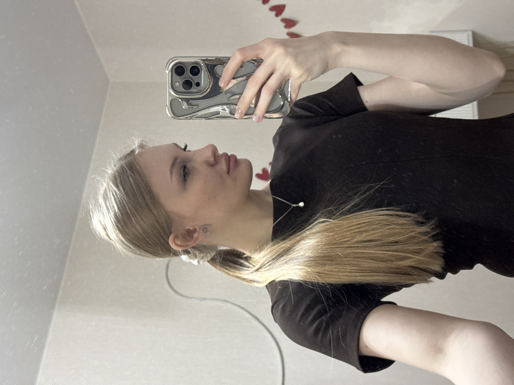 A young woman is taking a selfie in a room. She is holding a smartphone with a silver phone case in her right hand. She has light blonde hair pulled back into a low ponytail. She wears a black top and a delicate silver necklace. The background is a white wall with small red heart decorations. The location appears to be a private residence. It cannot be determined if the location is in Perm, Russia from the image provided.