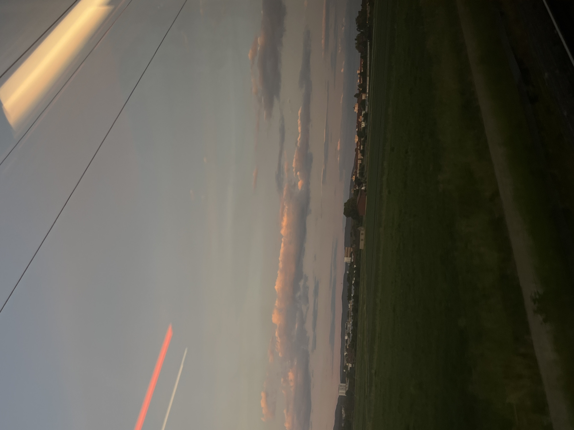 The image depicts a scenic view from a moving vehicle, likely a train, overlooking a rural landscape at dusk. The sky is a muted gradient of gray and pale pink, with scattered clouds tinged with the warm hues of sunset. A distinct contrail from an airplane is visible in the upper left portion of the sky. In the mid-ground, a line of buildings representing a town or village stretches across the horizon, with some structures appearing more prominent than others. A large, green field dominates the foreground, suggesting agricultural land. The lower right portion of the image shows a blurred reflection on the window, possibly from interior lights of the vehicle, and a dark strip suggesting a window frame or the edge of the vehicle. The overall impression is one of a serene, twilight journey through the countryside.