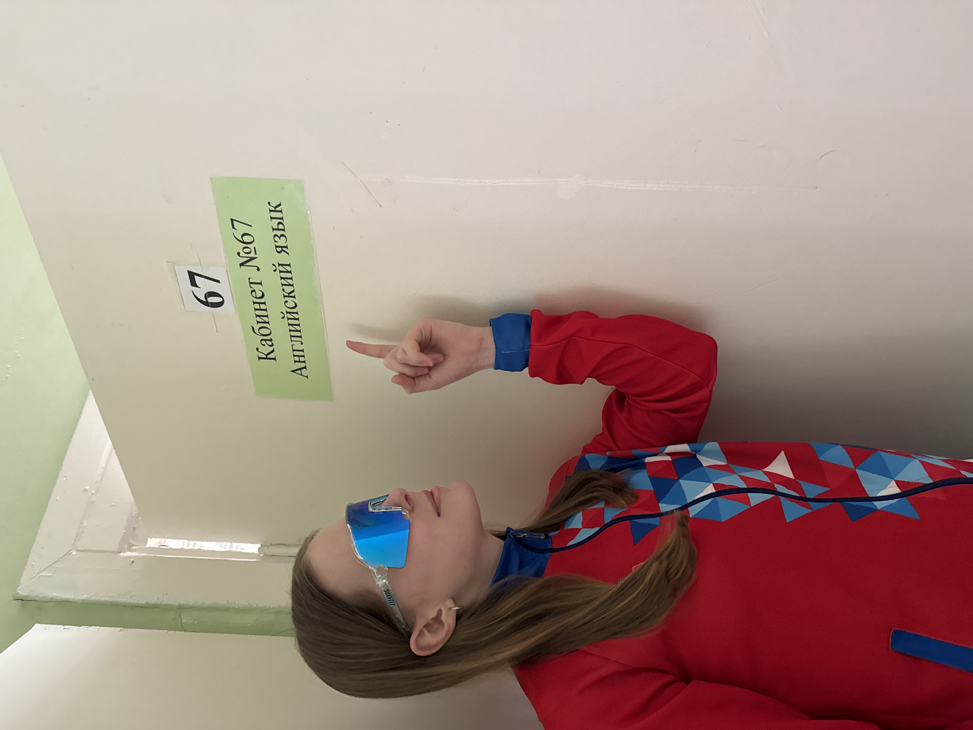 A young person with brown hair is pointing at a sign on a white wall. The sign is green and says "Кабинет №67 Английский язык" which translates to "Room 67 English Language." A smaller sign with "67" is placed on the wall above the larger sign. The person is wearing a red jacket with blue accents and blue sunglasses. The scene appears to be indoors, potentially in a school or building in Usolye, Russia, as indicated by the sign's language.