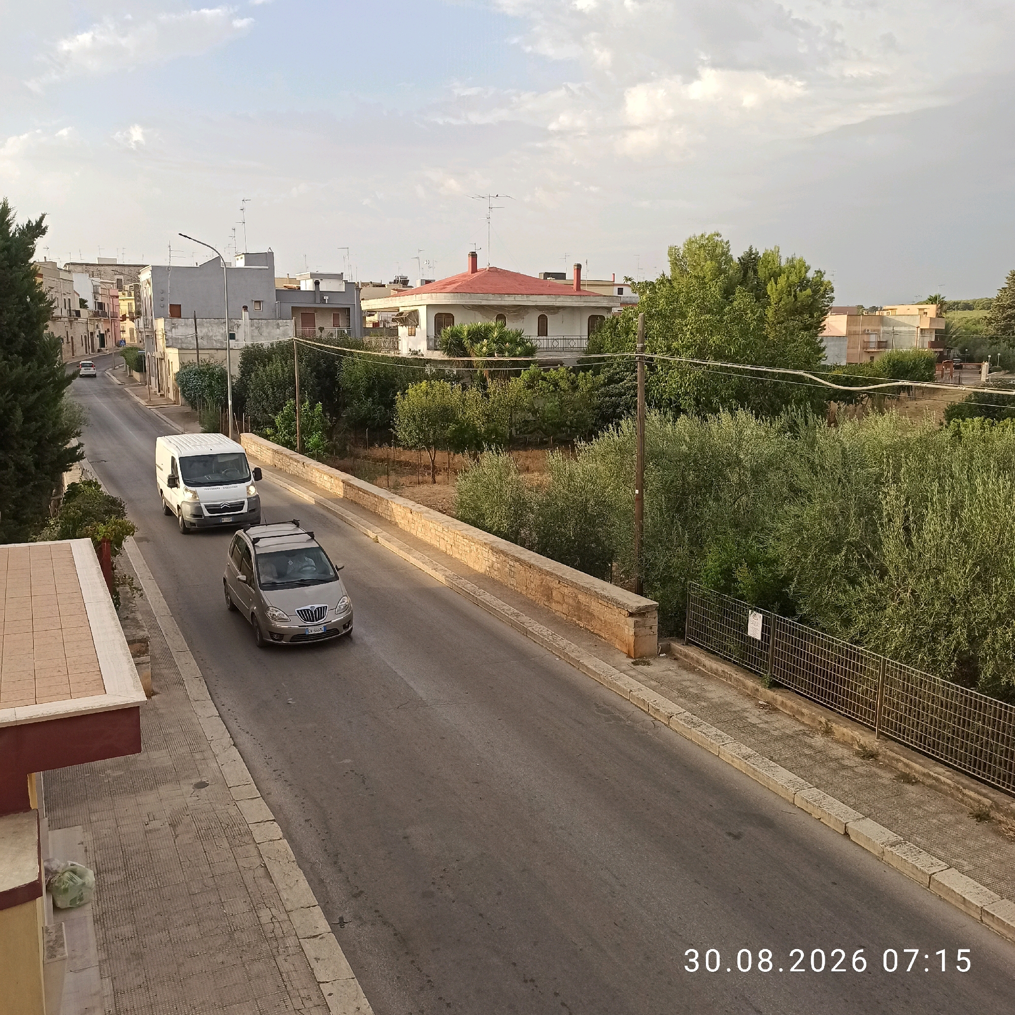 The image shows a street scene in Bitonto, Italy. A white van and a silver car are driving on the road. There are buildings and trees on either side of the street. The sky is cloudy. The date and time stamp at the bottom right corner of the image indicates it was taken on August 30, 2026, at 07:15. The overall scene depicts a quiet morning in a residential area.