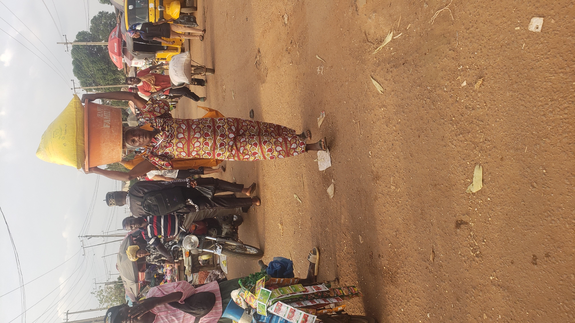 In Kakuri Gwari, Nigeria, a woman walks through a crowded outdoor market, carrying a large orange basin filled with a yellow sack on her head. She wears a vibrantly patterned traditional dress. To her left, a man in a black cap and traditional outfit, carrying a black backpack, walks past. Further left, a person in a pink striped shirt is partially visible. In the background, several people are present, some near stalls with umbrellas, and a yellow motorized tricycle and a motorcycle are visible. The ground is dirt.